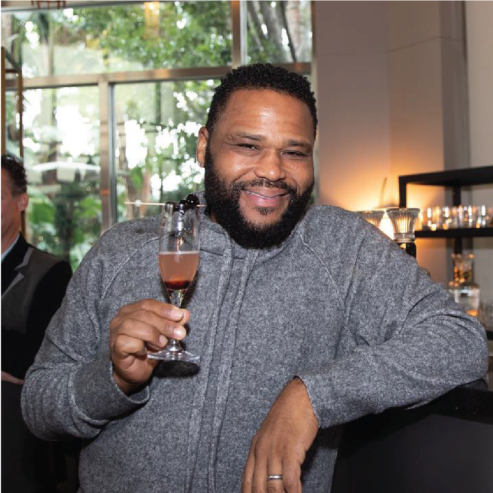 A smiling Anthony with a beard holding a glass of pink champagne, standing indoors near a bar, with a window and greenery in the background.