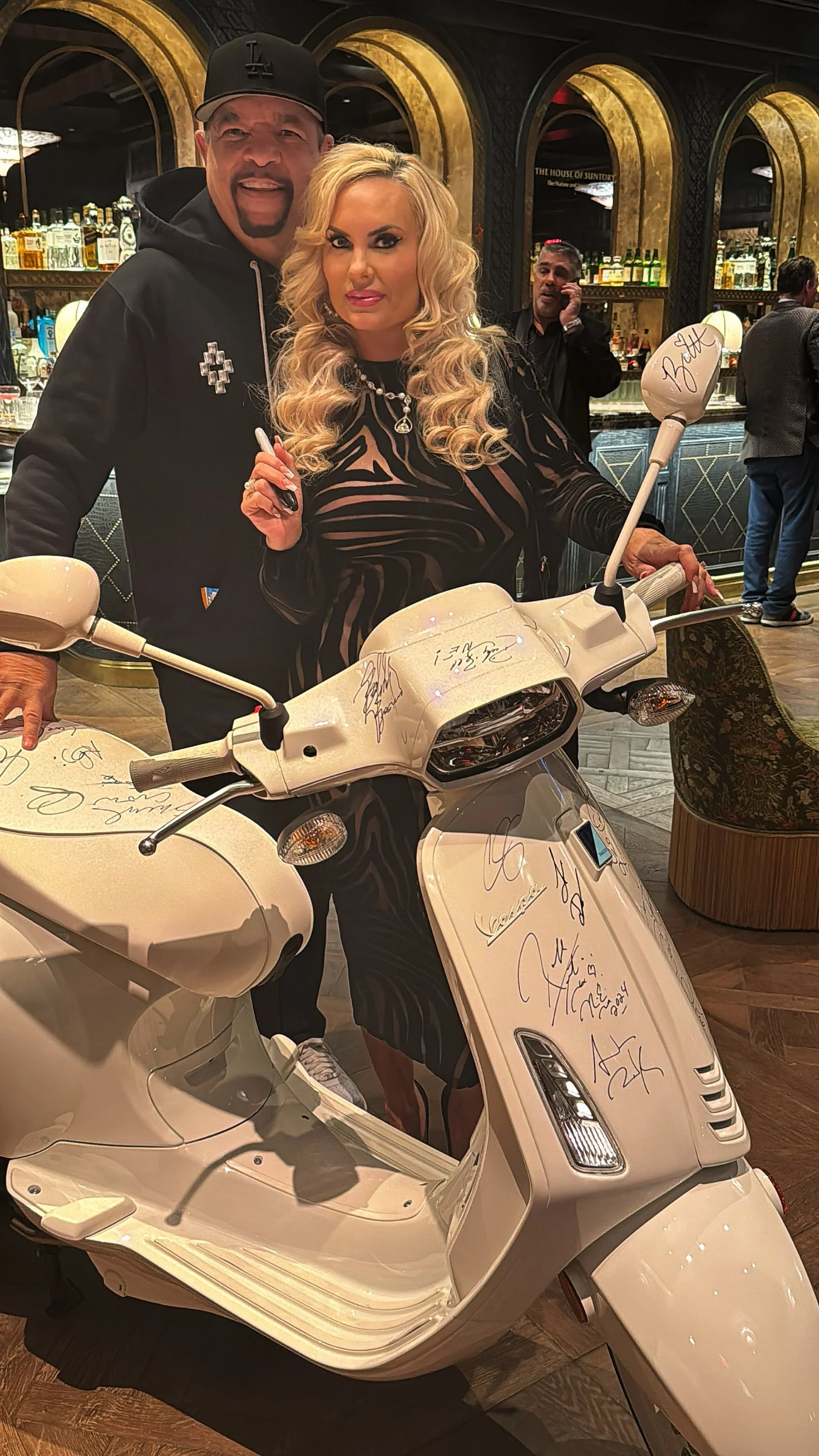 Woman with blonde curly hair in a black dress standing behind a white Vespa scooter with signatures and autographs, with two men in the background in a bar or lounge setting.