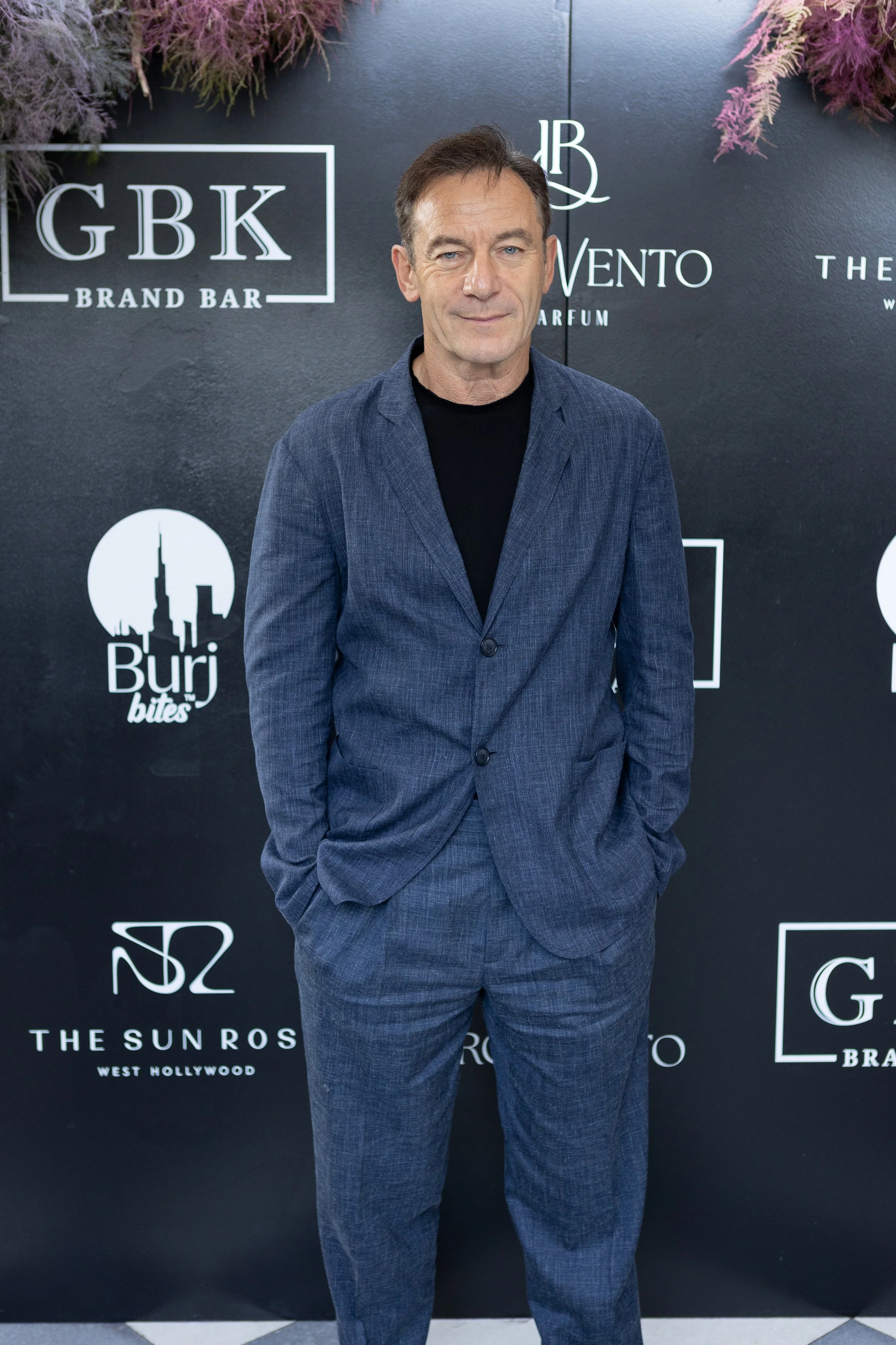 A man in a gray suit standing in front of a black backdrop with various logos and text, posing for a photo.