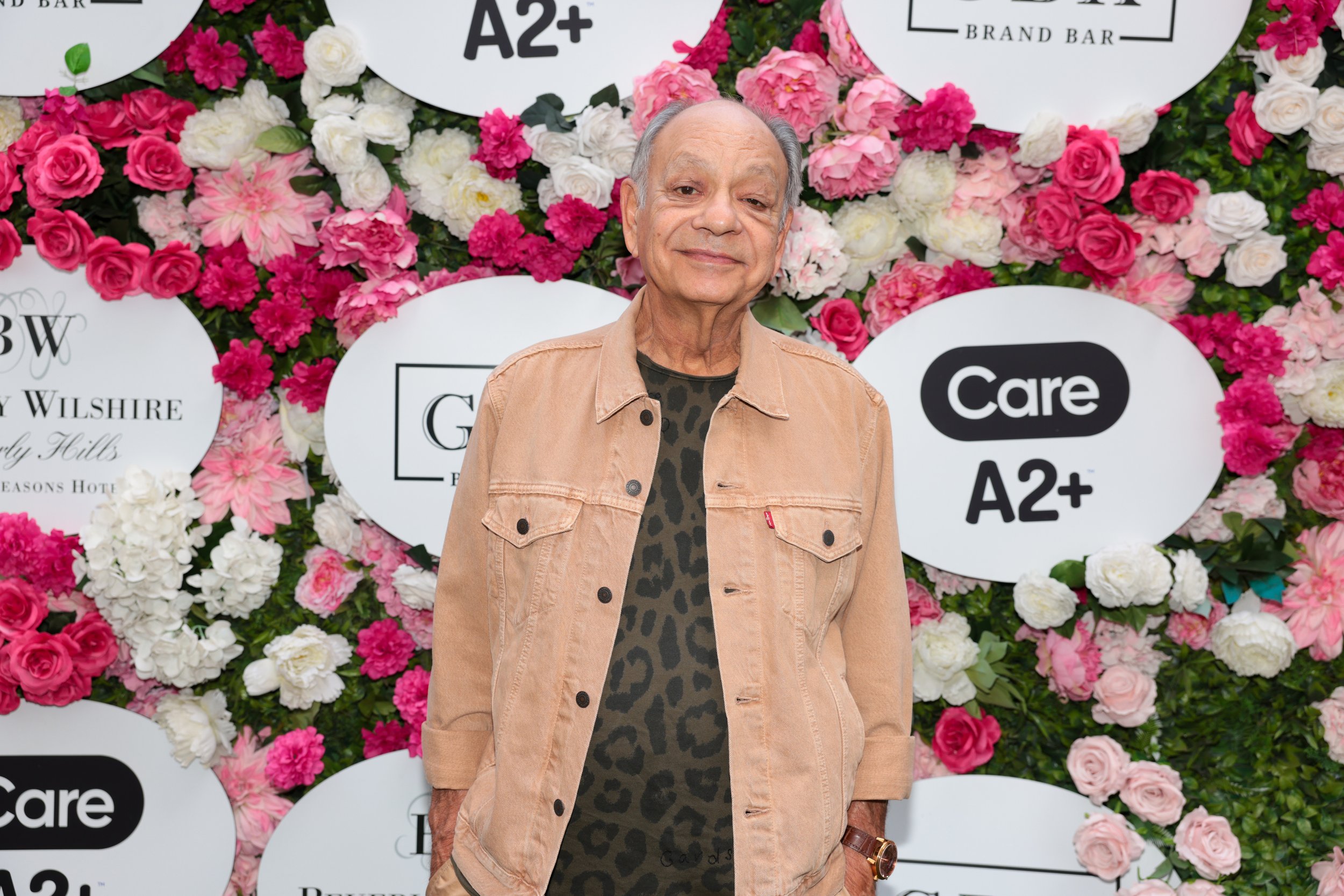 An elderly man in a beige denim jacket and leopard print shirt standing in front of a floral backdrop with white signs, including one that reads 'Care A2+'.
