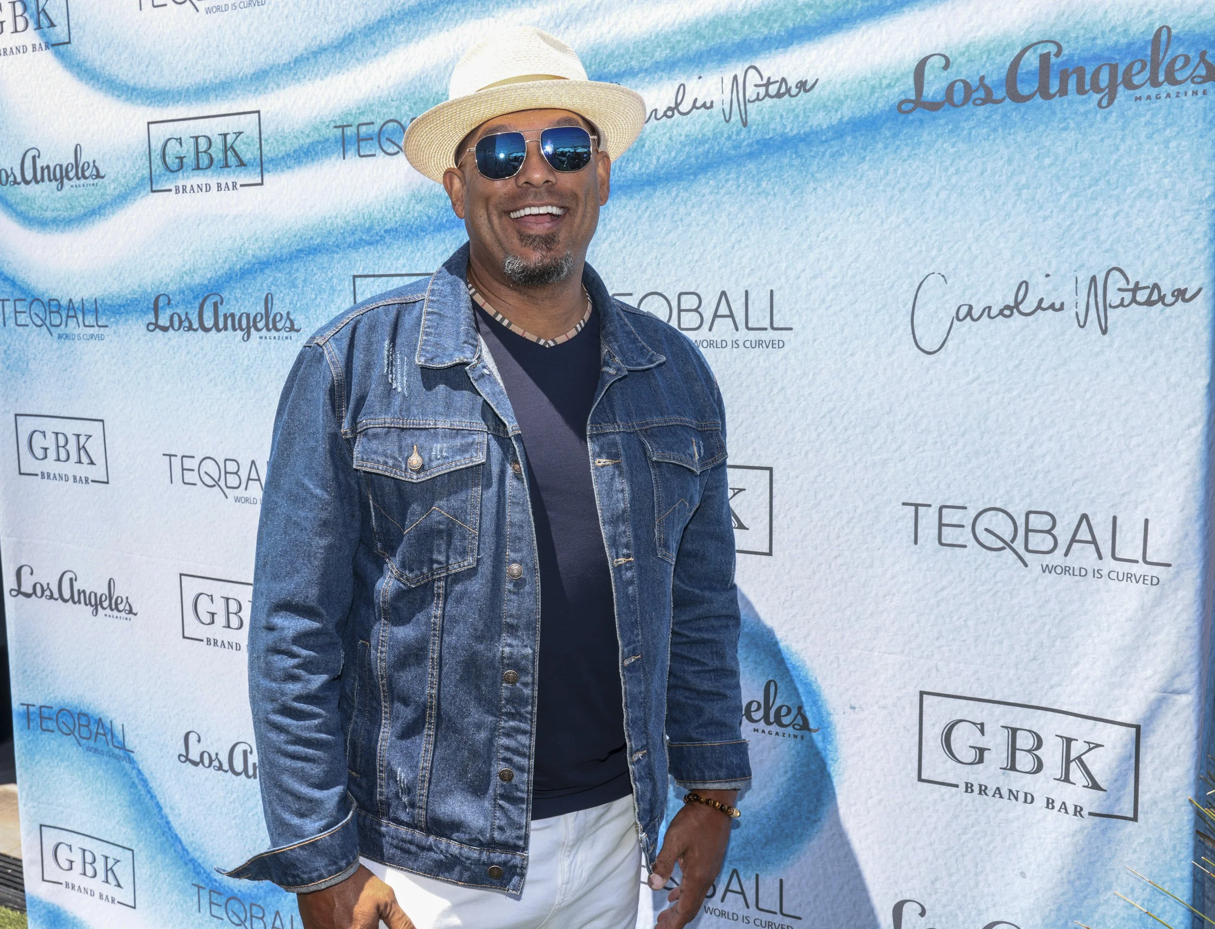 A man wearing a white hat, blue sunglasses, a denim jacket, and a dark shirt, smiling in front of a Los Angeles-themed backdrop with logos for GBK, Teqball, and Los Angeles Magazine.