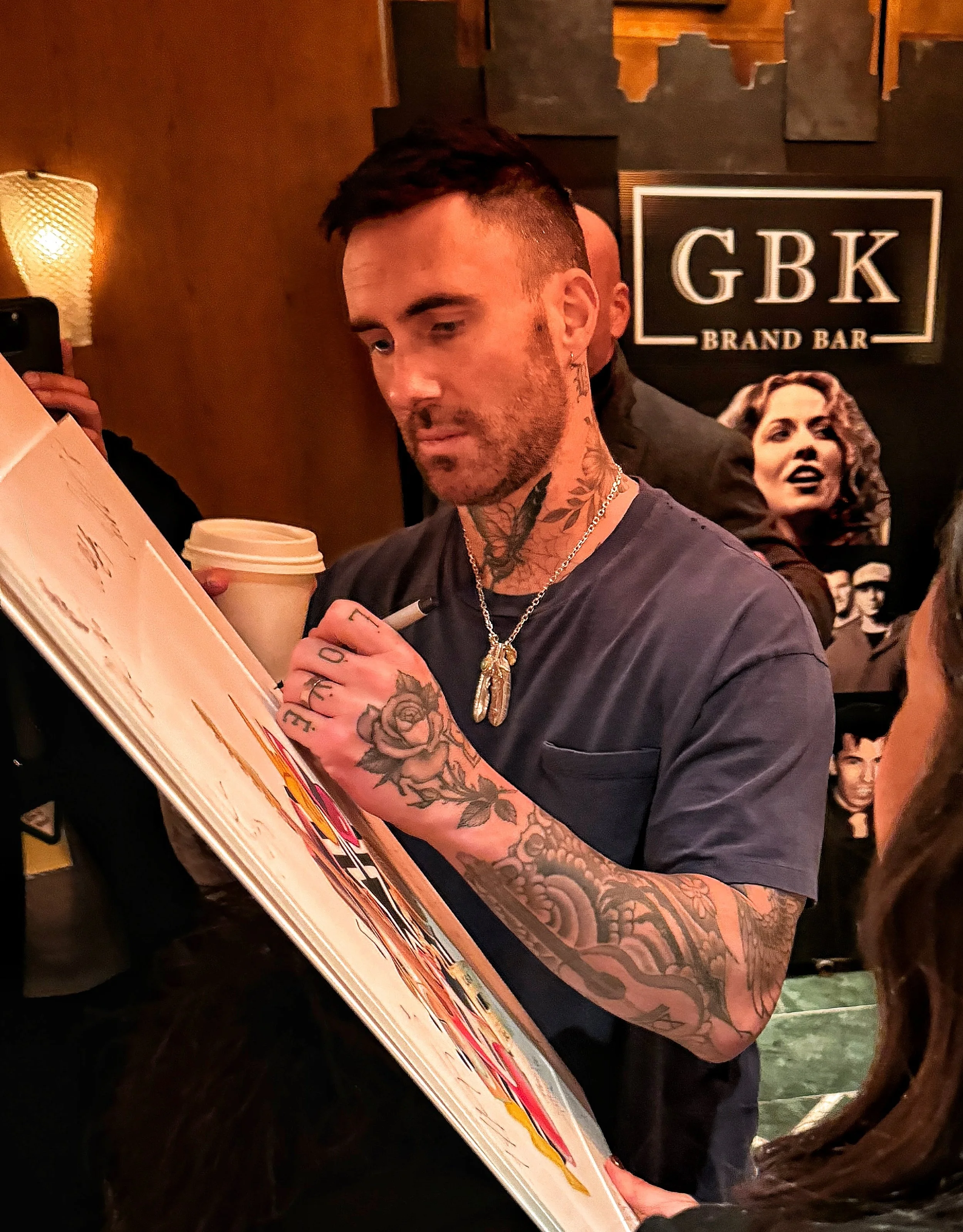 A man with tattoos on his neck and arms signing an autograph at a bar named GBK Brand Bar, holding a coffee cup, with a background poster of women and people.