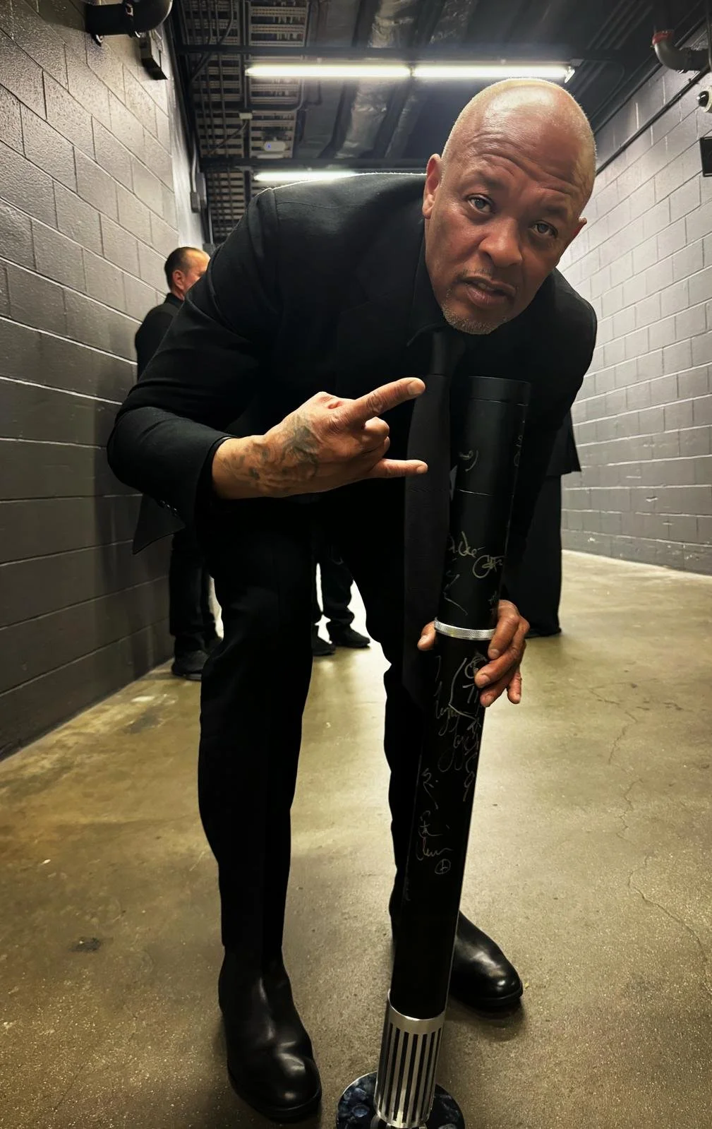 Bald man in black suit leaning forward and making a rock and roll hand sign, holding a black microphone with signatures, in a gray hallway with another man in black in the background.