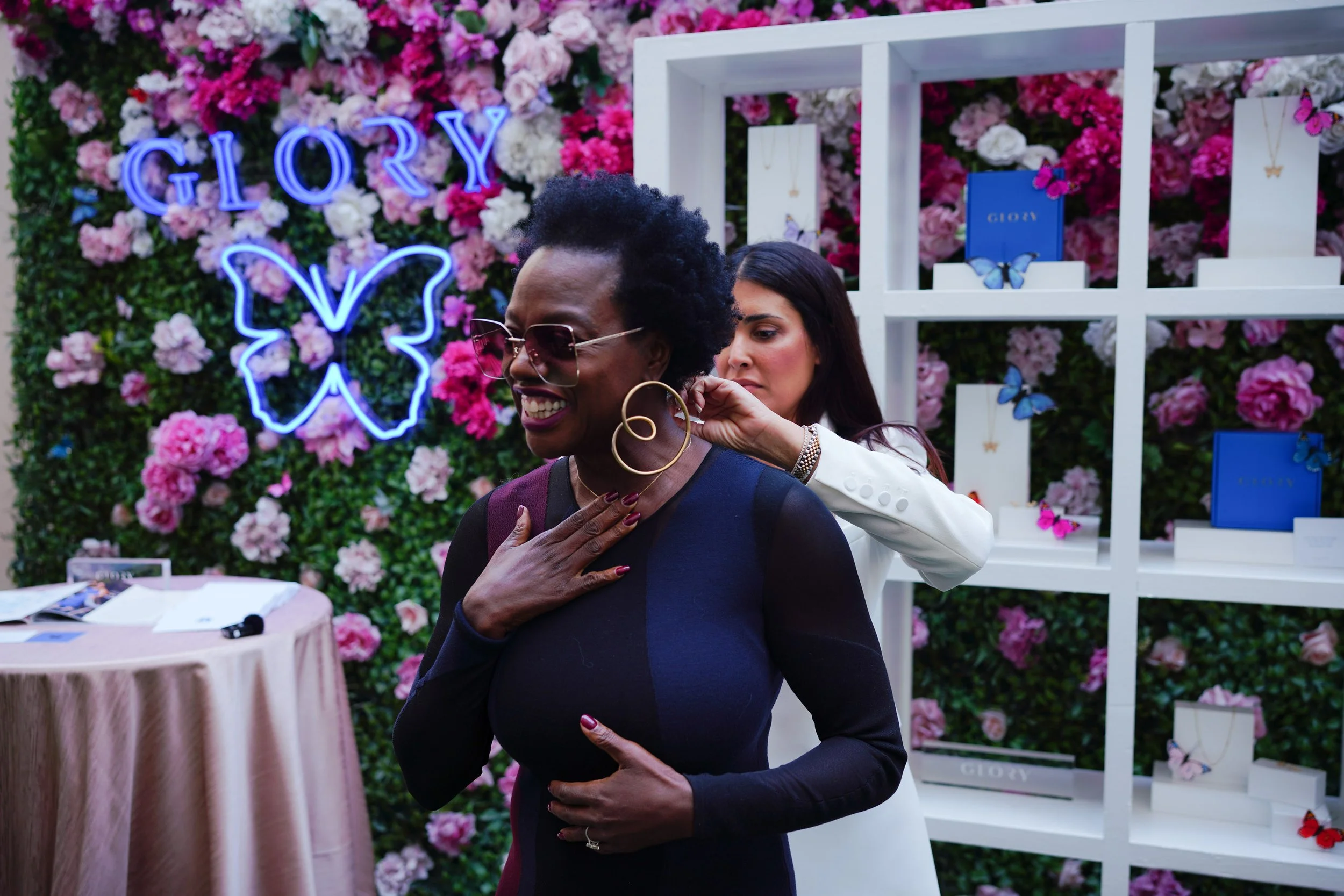 Two women at a jewelry booth with a floral backdrop and butterfly decorations, one trying on jewelry.