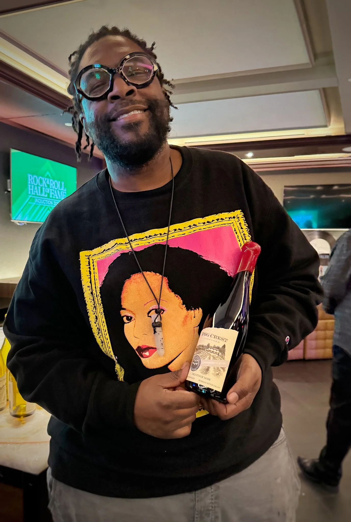 Man with glasses smiling, wearing a black sweatshirt with a colorful graphic of a woman's face, holding a bottle of wine labeled fog crest in a social setting.