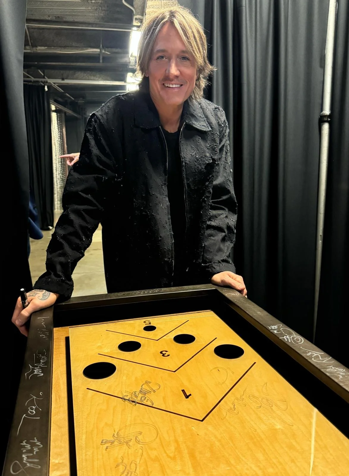 A man with shoulder-length blonde hair smiling and standing behind a vintage air hockey table in a dimly lit room with black curtains. He is wearing a black jacket and holding a paddle in his right hand.