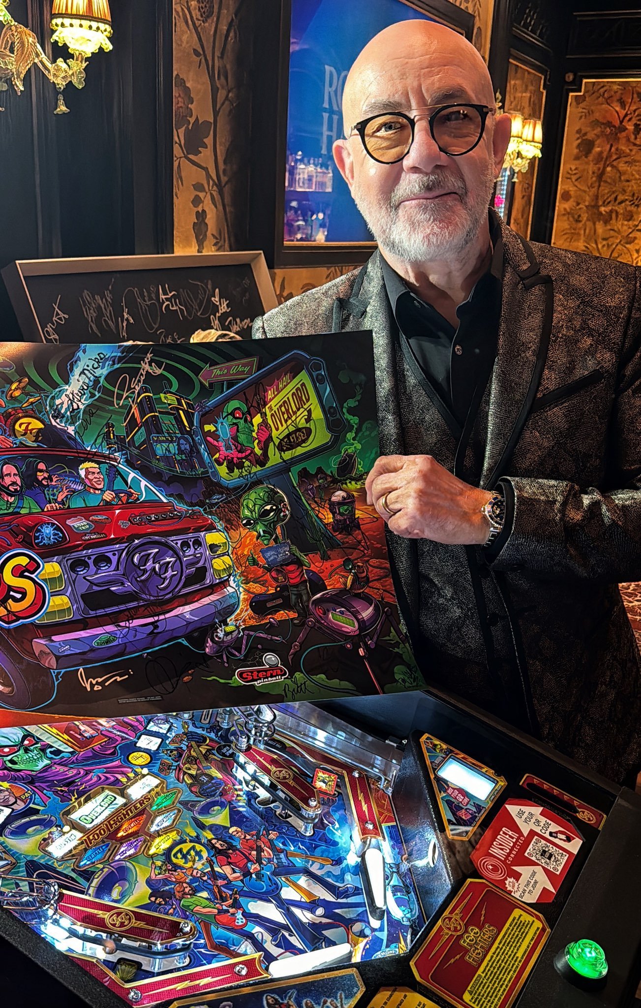 A man with glasses and a beard, dressed in a dark jacket, holding a colorful pinball machine artwork in a dimly lit, vintage-style room with ornate wallpaper, lamps, and framed pictures.