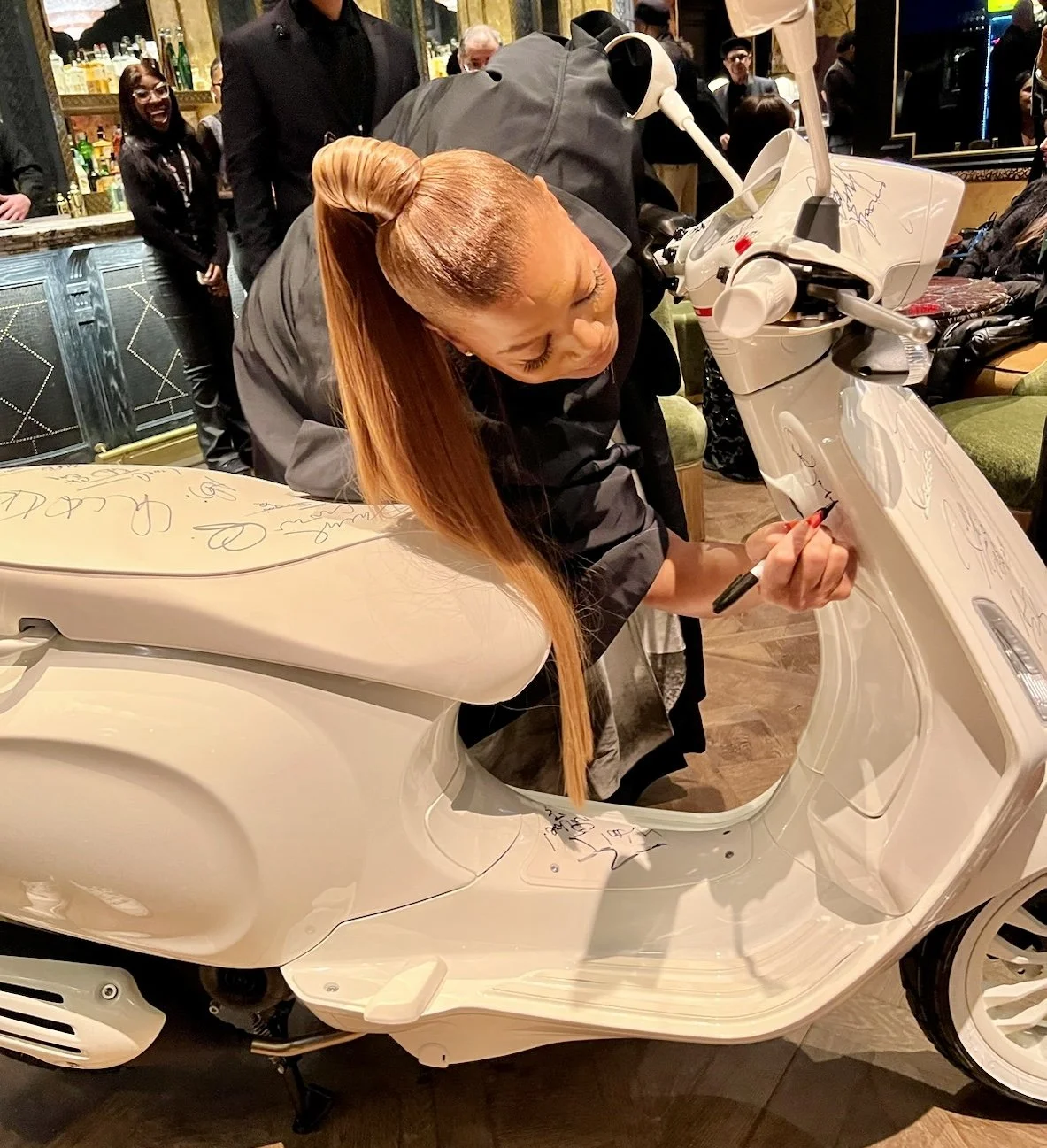 A woman with long red hair signing a white scooter while bending over it, surrounded by people in a dimly lit indoor setting, with some of them smiling and watching.