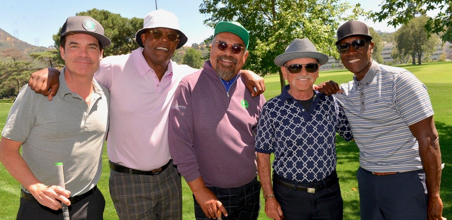 Five men standing together on a golf course, smiling, with trees and hills in the background.