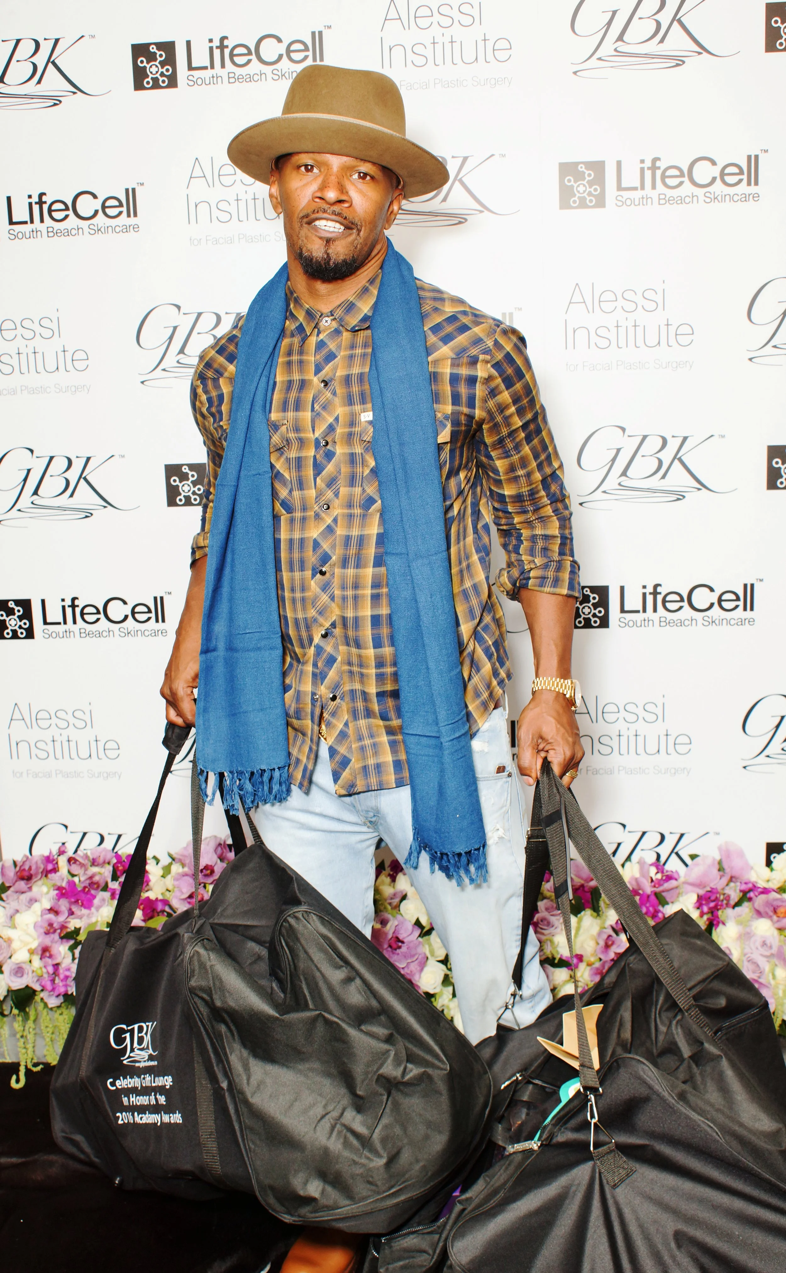 A man dressed in a plaid shirt, light jeans, a wide-brimmed hat, and a blue scarf, standing on a stage with a backdrop featuring logos for GBK, LifeCell, and Alessi Institute. He is holding large black bags and standing next to a floral arrangement.