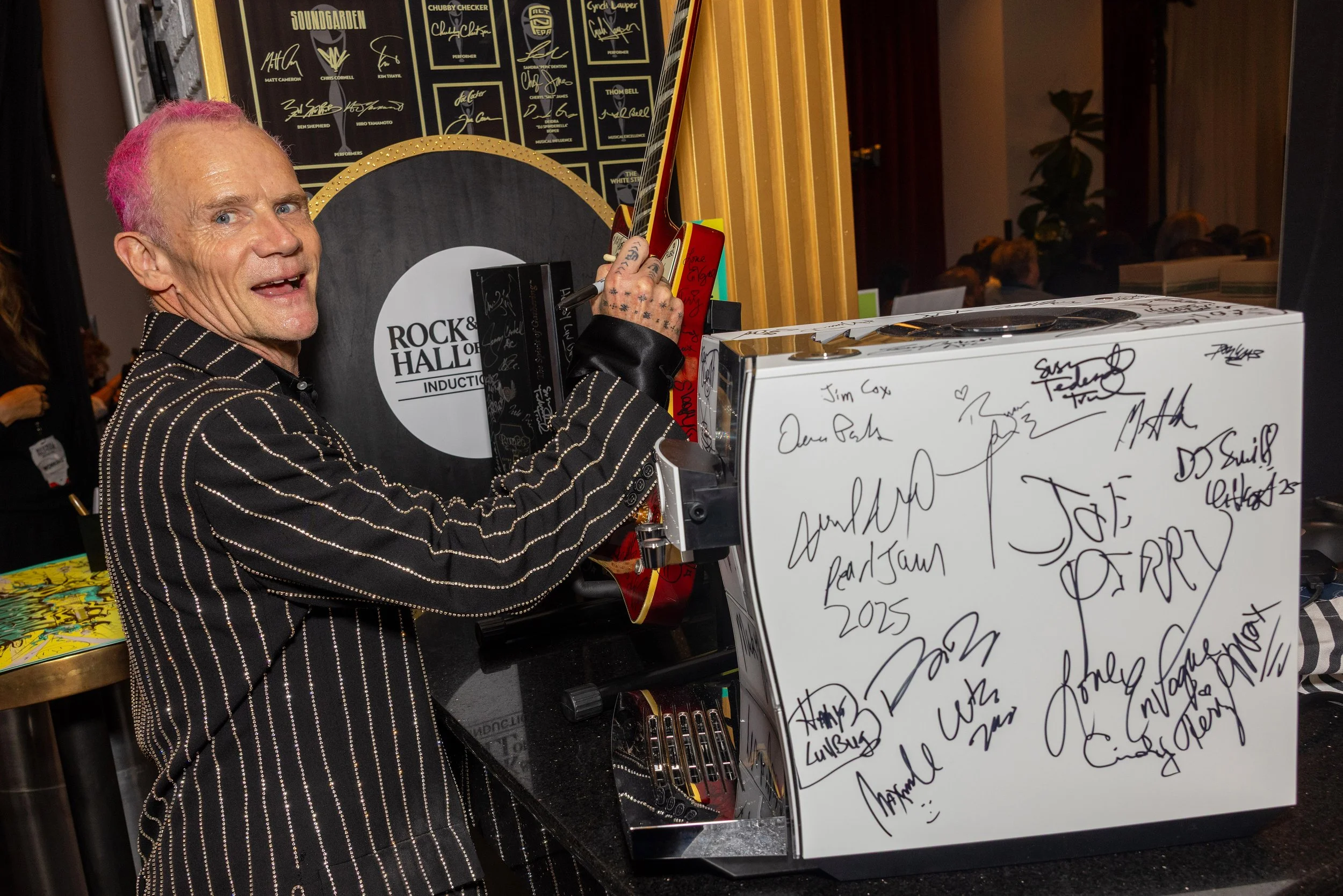 A man with pink hair signing autographs at a table with a white board covered in signatures and messages. Behind him are posters and a guitar on display.
