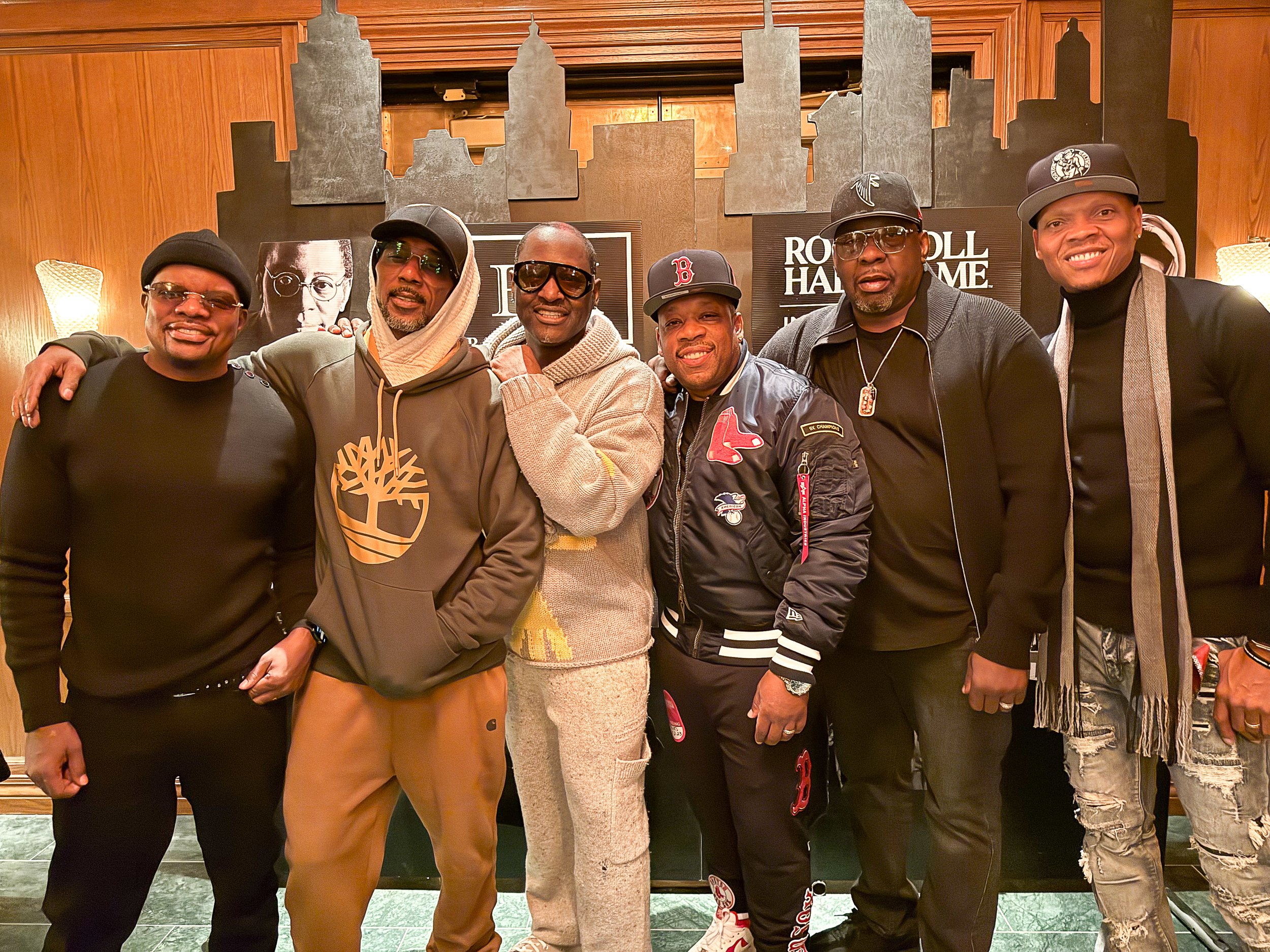 Seven men posing together, smiling, indoors with wooden accents and city skyline cutouts in the background.