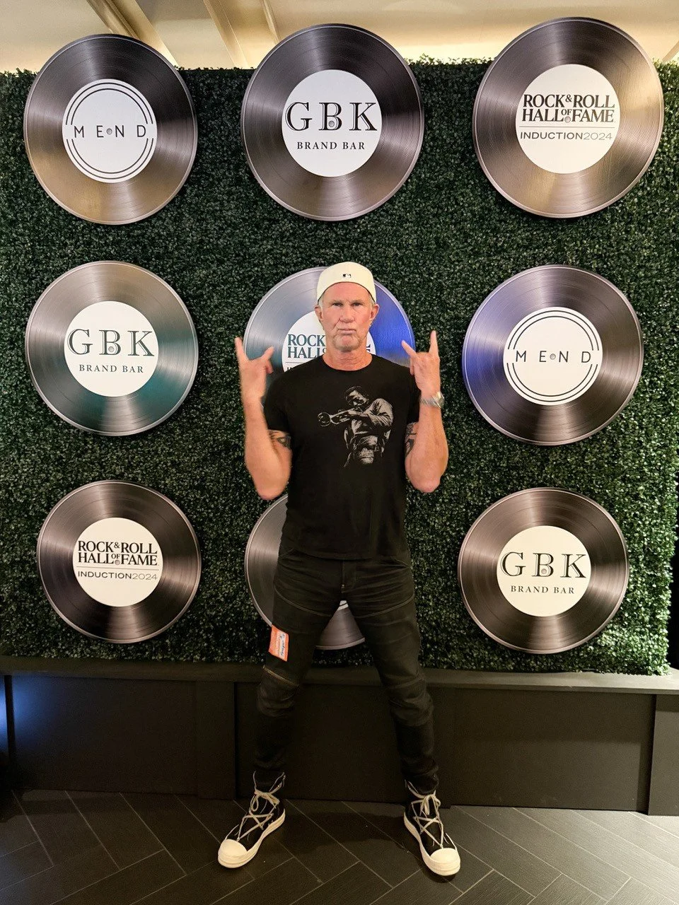 A man wearing a black T-shirt with a graphic, black pants, and white sneakers standing in front of a decorative wall with eight circular metallic plaques, each featuring different logos or text, including 'MEND', 'GBK Brand Bar', and 'Rock & Roll Hall of Fame Induction 2024'. The man is making a rock gesture with both hands and looking at the camera.