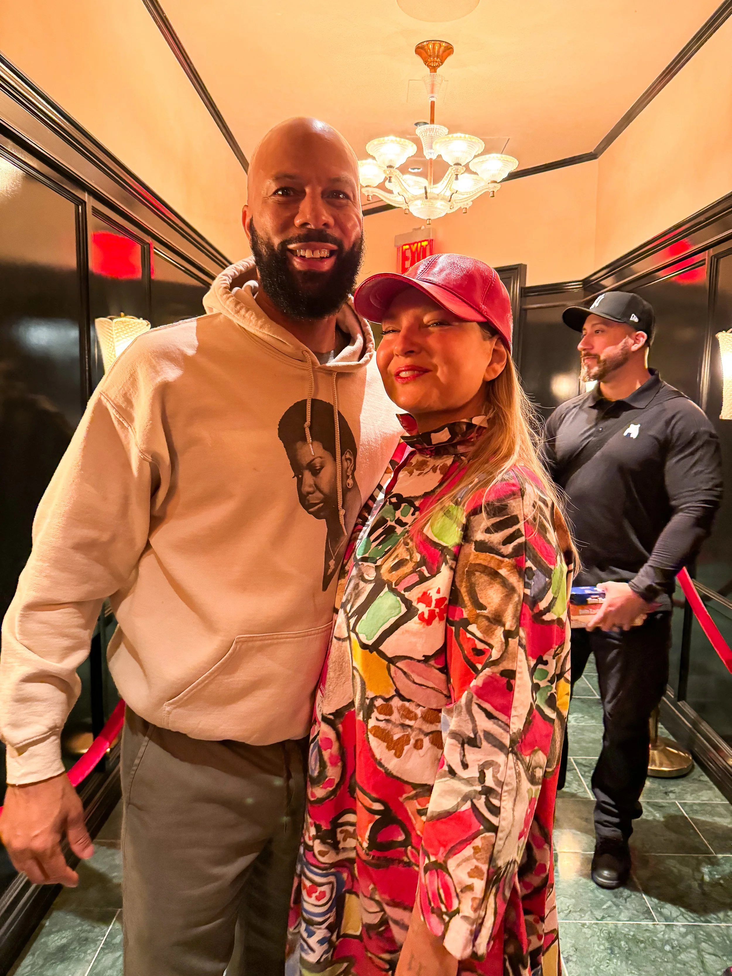 A smiling man with a beard wearing a beige hoodie with a woman in a colorful patterned jacket and red cap in a dimly lit hallway with black walls and chandeliers, with a doorman in black in the background.