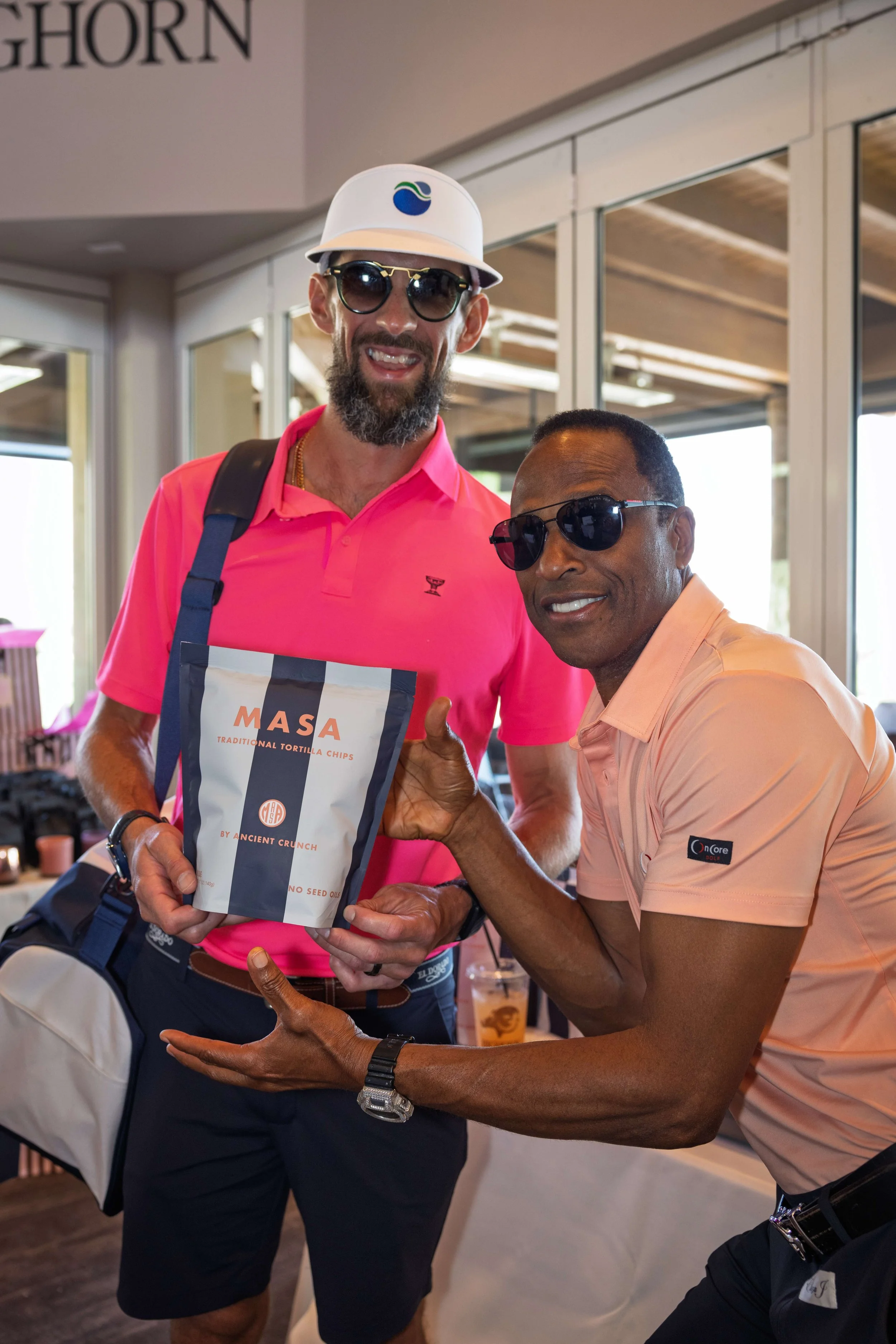 Two men wearing sunglasses and smiling, one holding a bag of MASA tortilla chips, posing indoors.