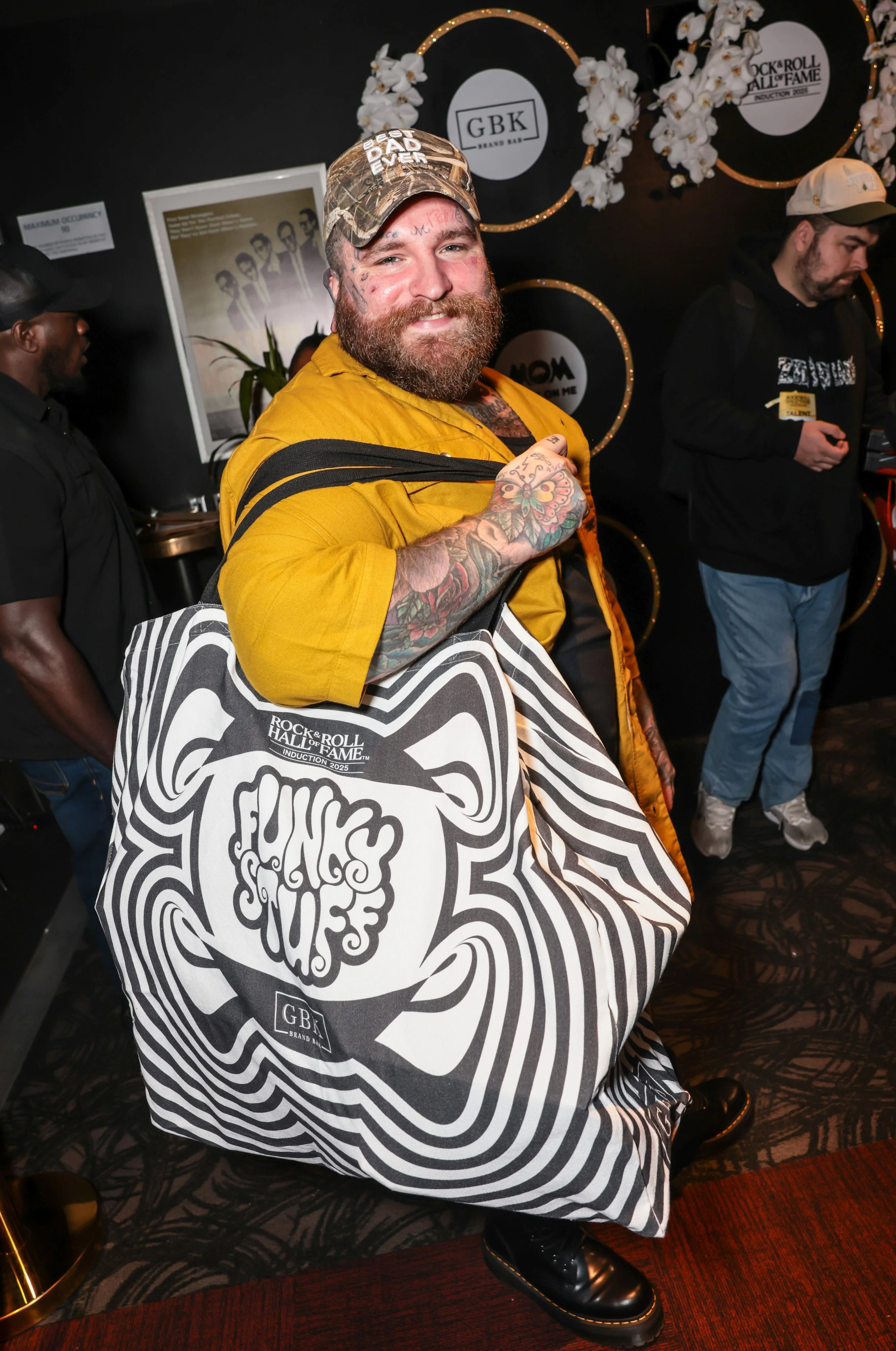 Man with tattoos, beard, yellow shirt, cap, and bag smiling at a music hall of fame event, with others in the background.
