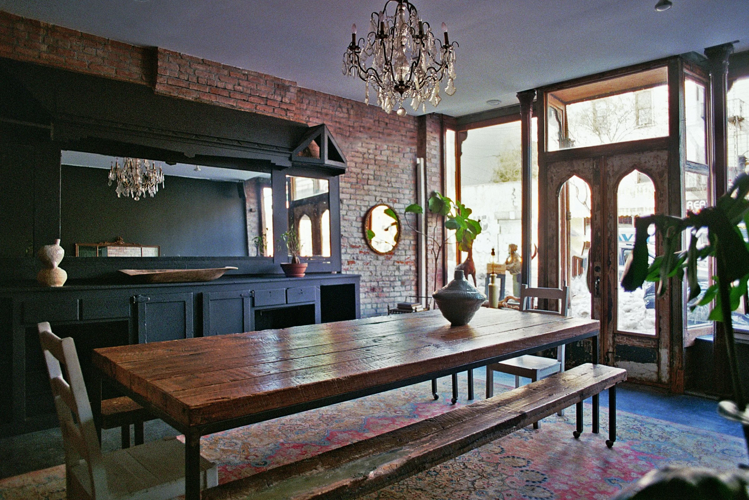Interior of a rustic cafe with a large wooden table, black sideboard with glassware and decorative vases, large mirror with moss-covered frame, vintage wooden doors, chandelier, brick wall, plants, and large windows letting in natural light.