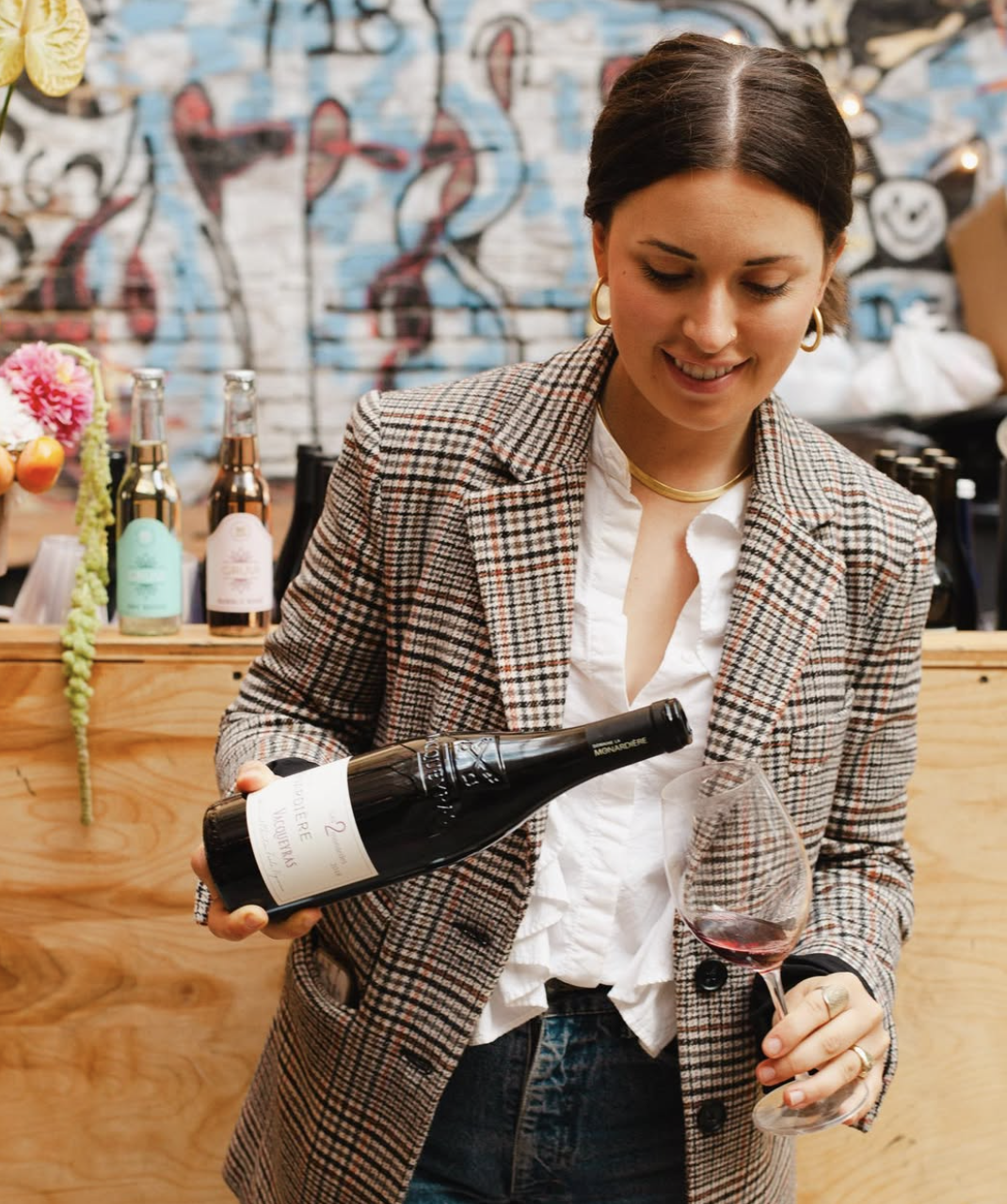 A woman in a plaid blazer pouring red wine into a glass at a bar with graffiti artwork on the wall.