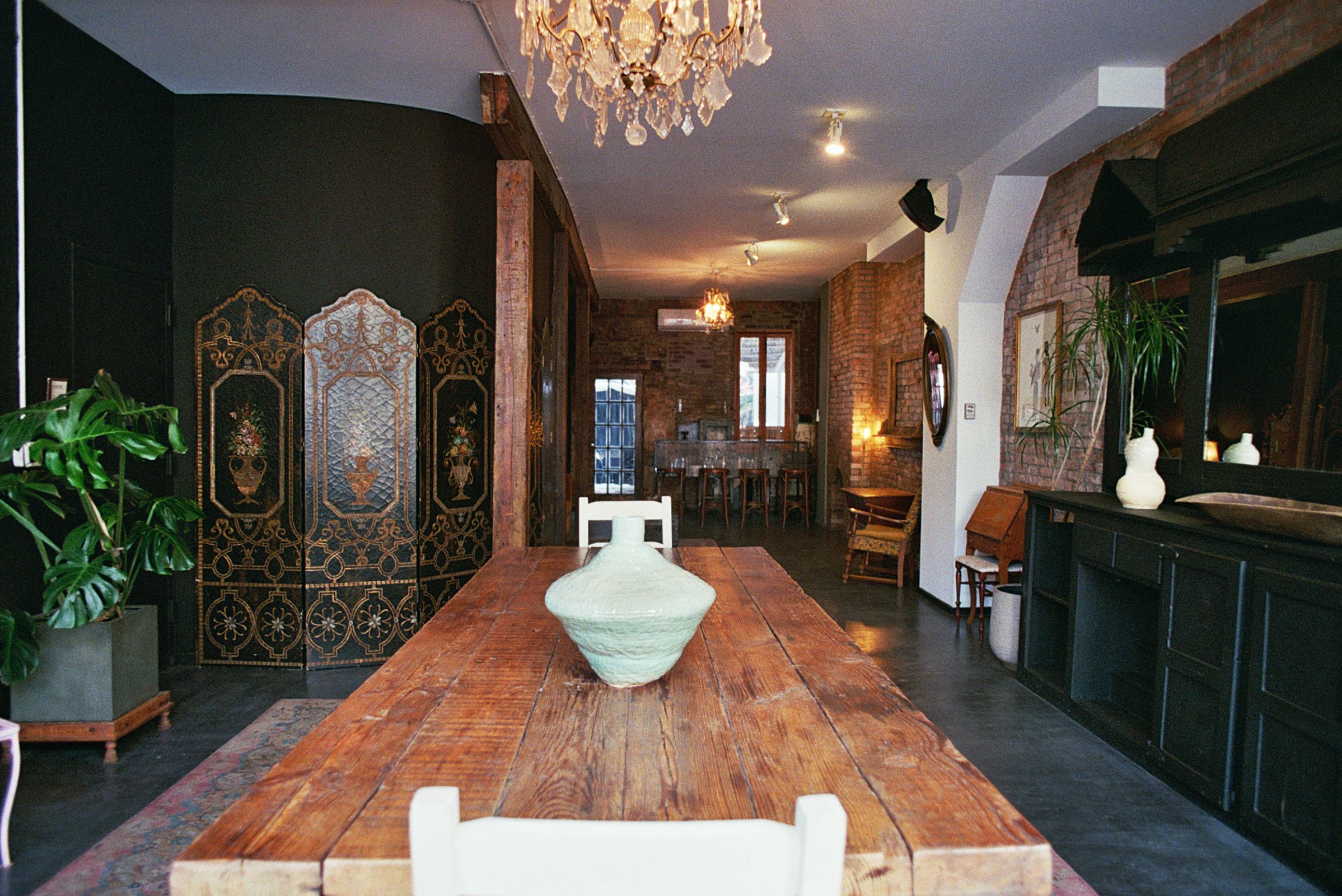 Interior of a stylish living room with exposed brick walls, a wooden dining table with a white vase and dried flowers, large green leafy plants in the foreground, a chandelier hanging from the ceiling, and a mirror with antler decoration above a black sideboard.