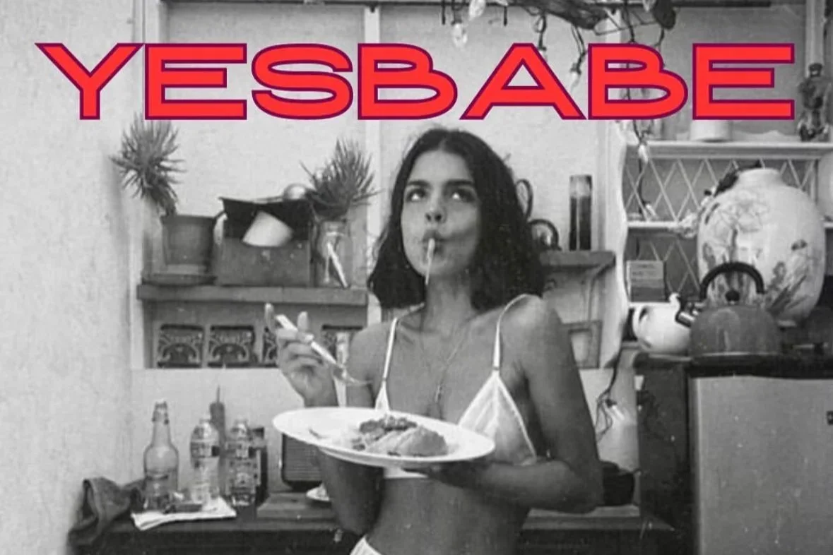 Black and white photo of a woman with short curly hair eating spaghetti with a fork, holding a plate and a cigarette, in a cluttered kitchen or living space. Large red text at the top reads "YESBÁBE".