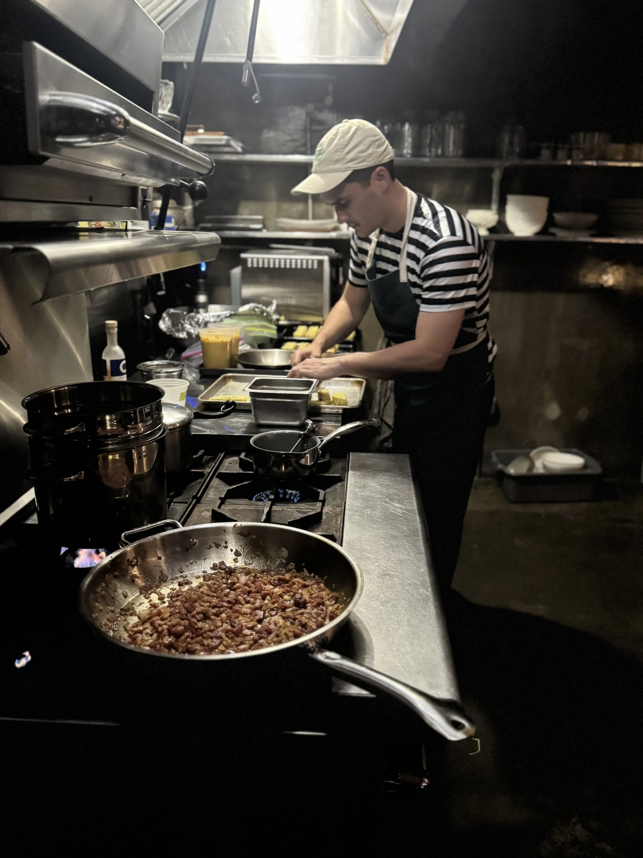 A chef cooking in a professional kitchen, with a front view of a pan containing browned chopped garlic and onions on a stovetop. The chef is preparing food at a countertop with various ingredients and utensils around.