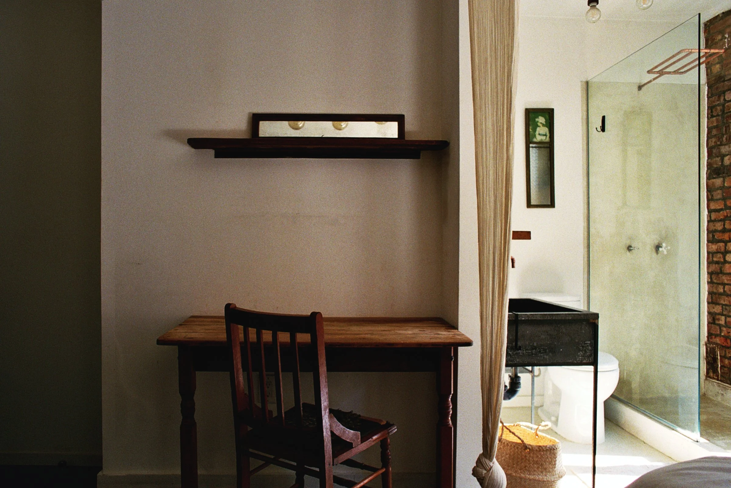 Interior of a cozy cafe with an exposed brick wall, a small round table with a bottle of wine, a glass, an open magazine, and assorted chairs. There is a vintage trunk and a red armchair nearby, with wall-mounted shelves and a picture frame.