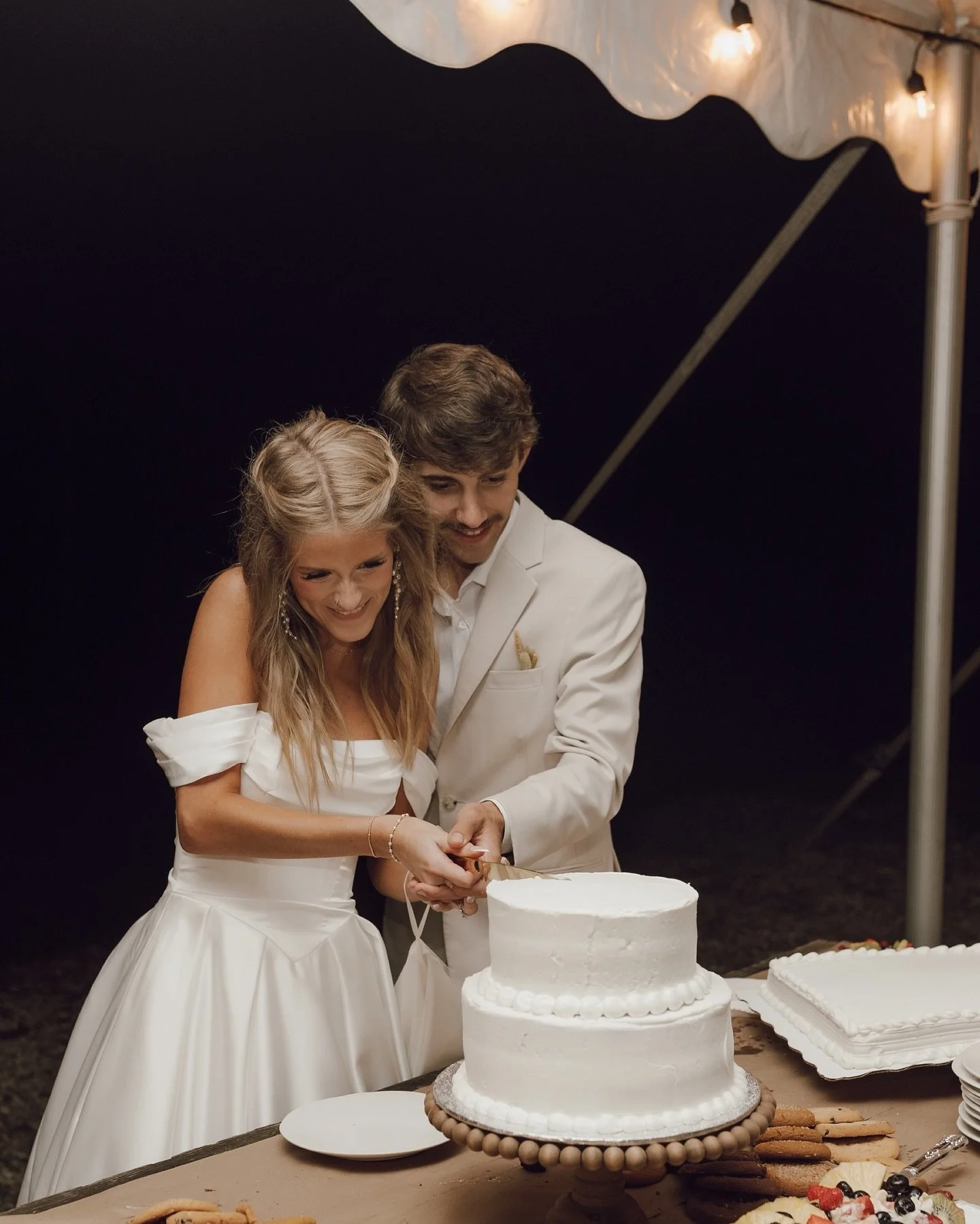 A bride and groom in white tuxedo and dress cutting a wedding cake together at night under a tent.