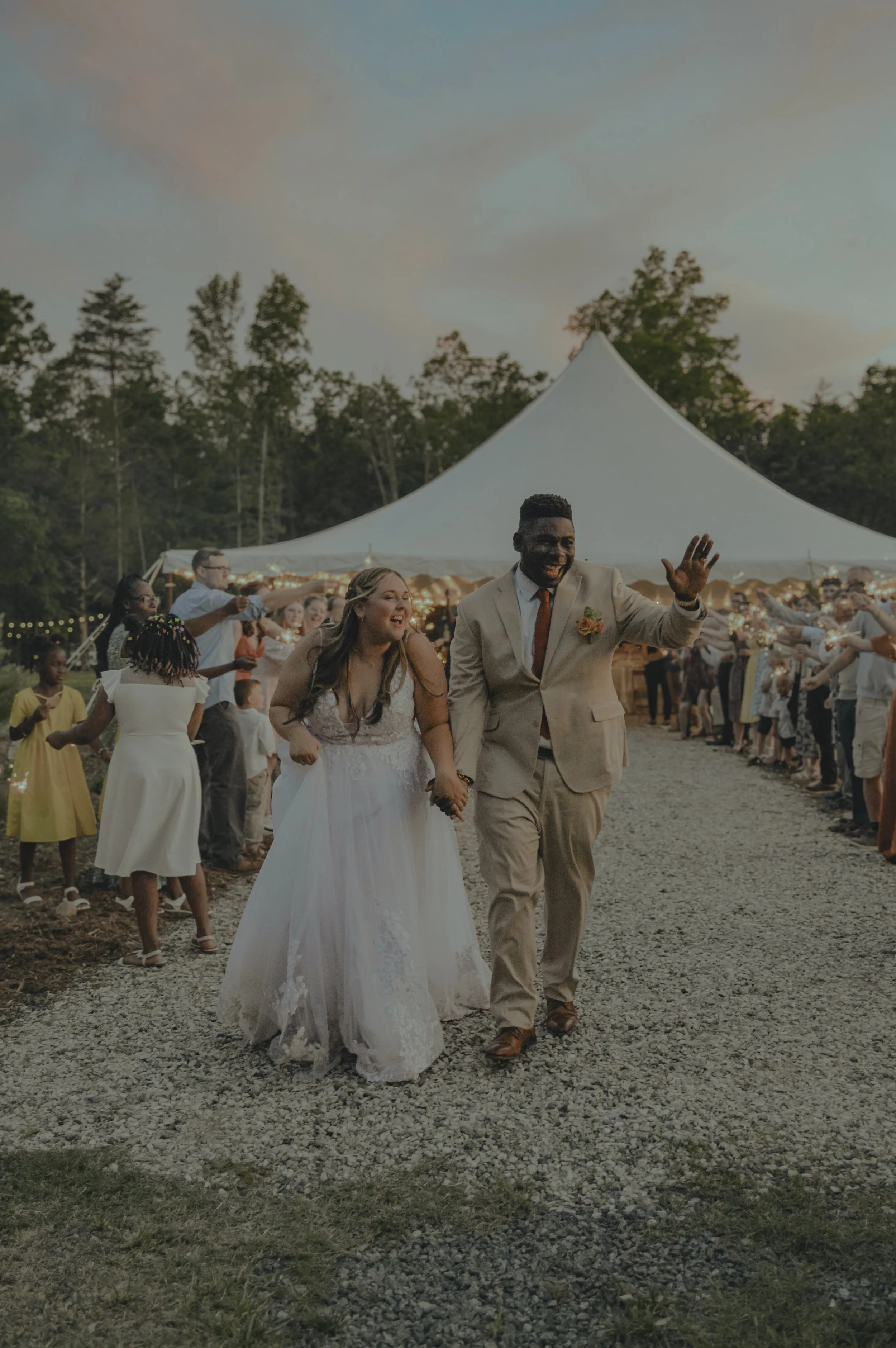 Bride and groom walking hand-in-hand under a large white event tent, surrounded by friends and family celebrating outdoors at sunset.