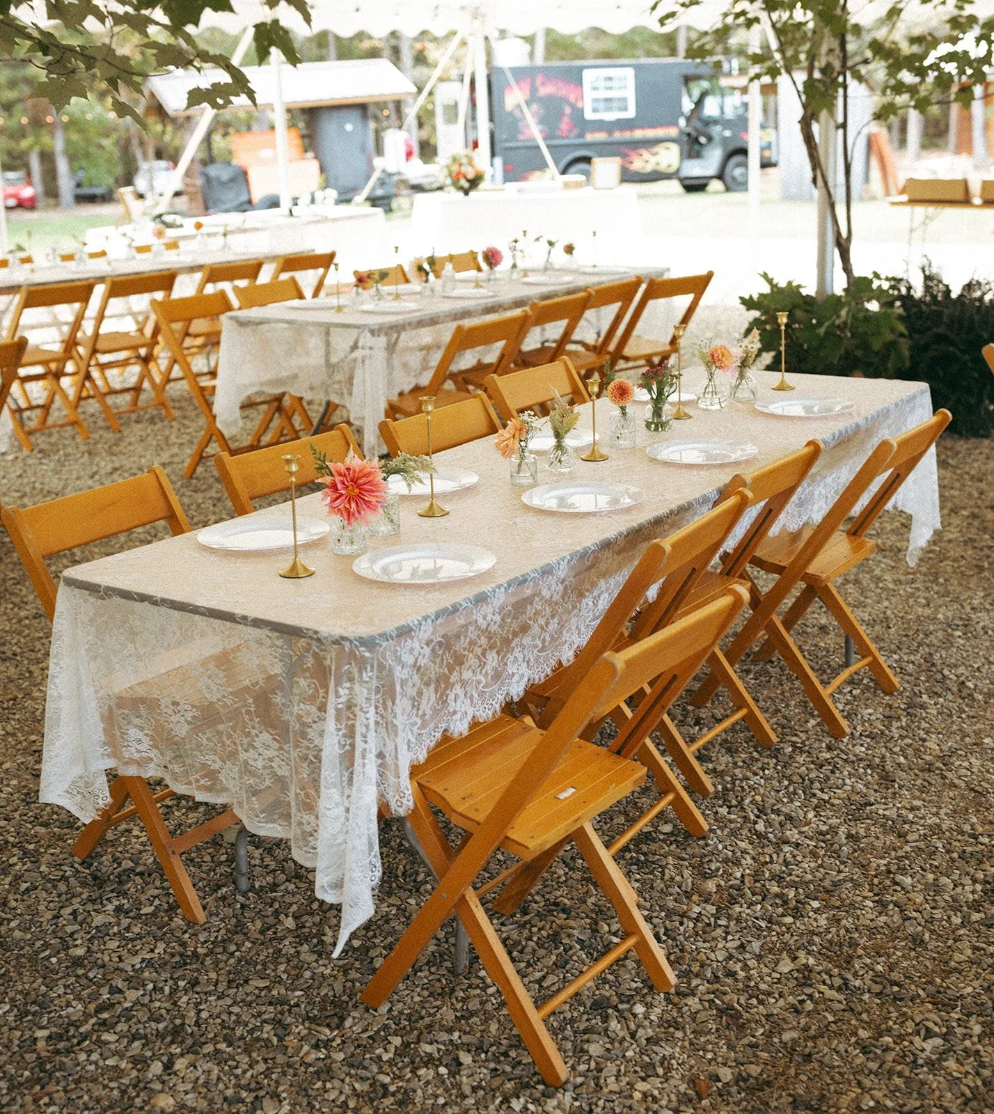 Set dining tables decorated with floral centerpieces and white lace tablecloths in an outdoor event space, with a food truck and trees in the background.