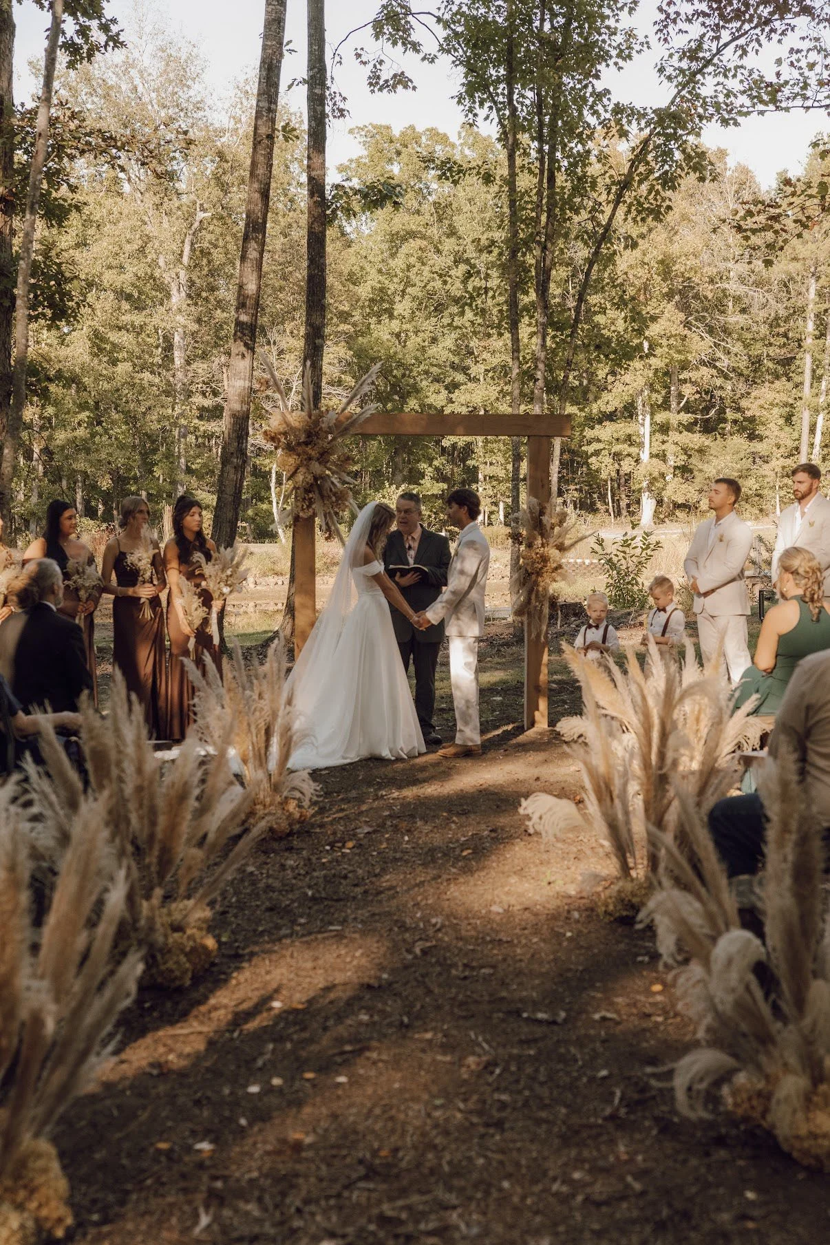 A wedding ceremony taking place outdoors in a forest, with the bride and groom holding hands and exchanging vows beneath a wooden arch decorated with dried flowers, surrounded by bridesmaids, groomsmen, and guests.