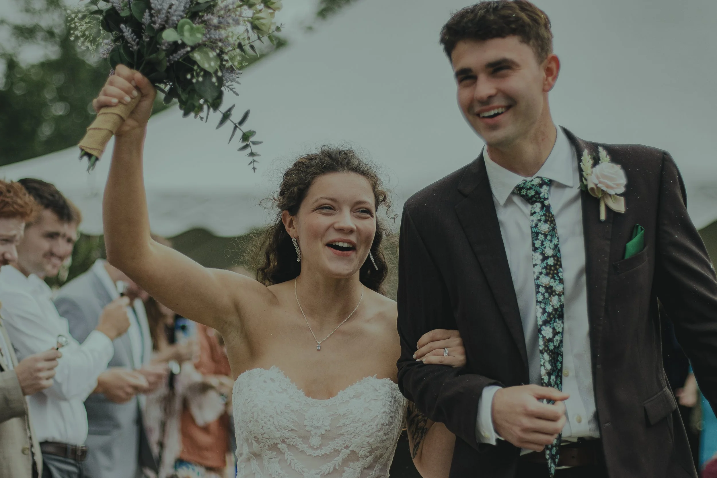 A bride and groom smiling at an outdoor wedding ceremony, with the bride holding a bouquet. Guests are visible in the background.