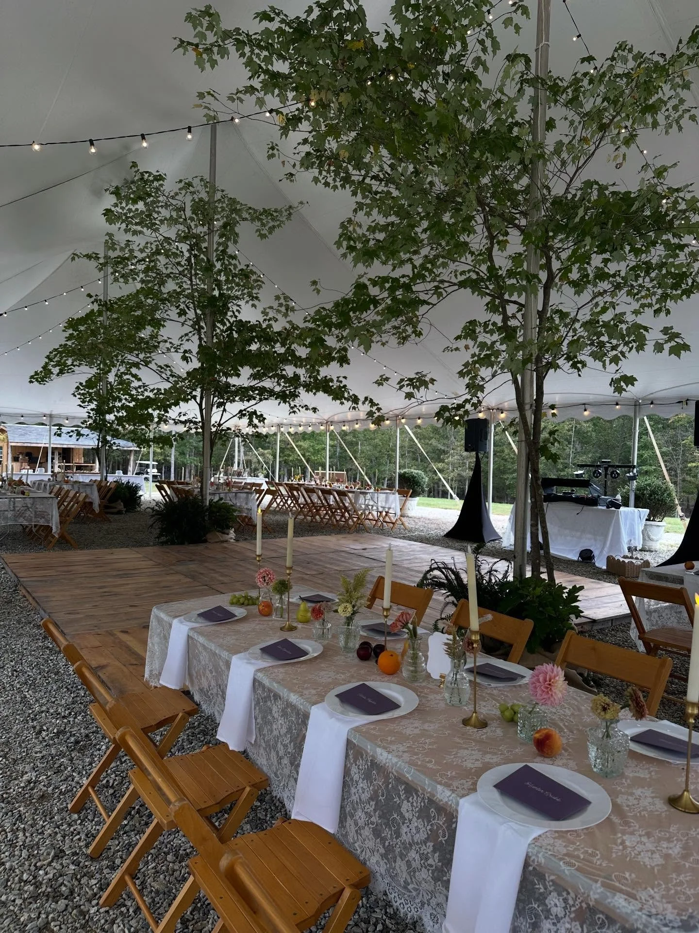 Under a large tent, a decorated event space features a long table with a lace tablecloth, set with plates, candles, flowers, and small fruit decorations. Trees with string lights are visible inside the tent, and additional tables and a DJ setup are s