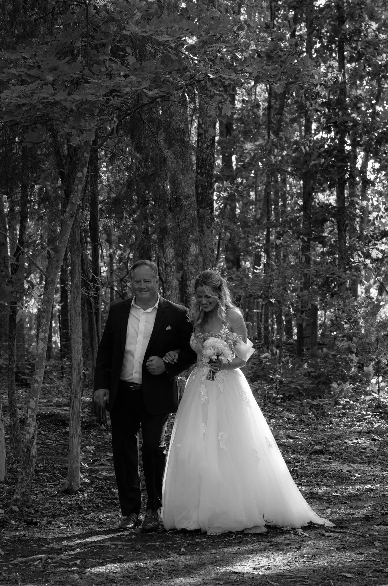 Black and white photo of a bride in a wedding dress and a man, possibly her father, walking arm in arm through a forested area.