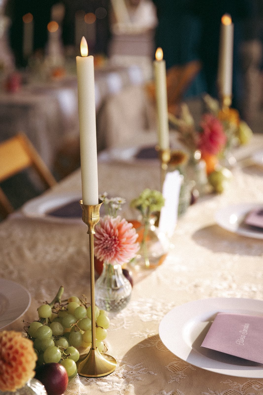 A close-up of a decorated dining table with lit white candles in brass holders, floral arrangements with pink and white flowers, and a bunch of green grapes, set for a formal event or celebration.