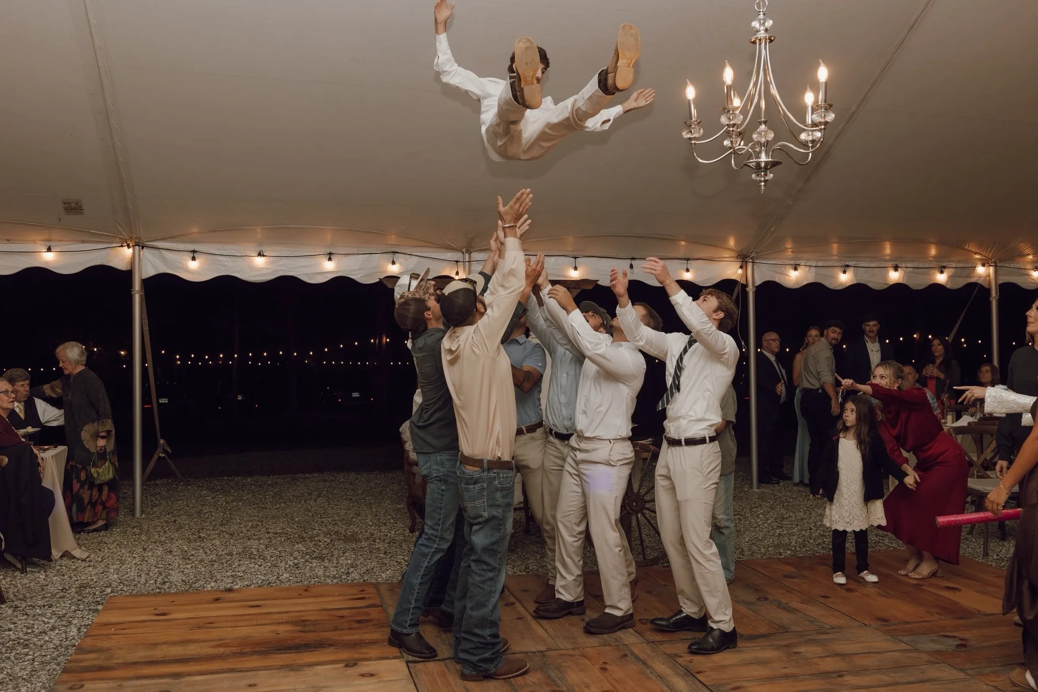 People celebrating at an outdoor event under a large tent, with one person being tossed into the air by a group, near a chandelier and string lights at night.