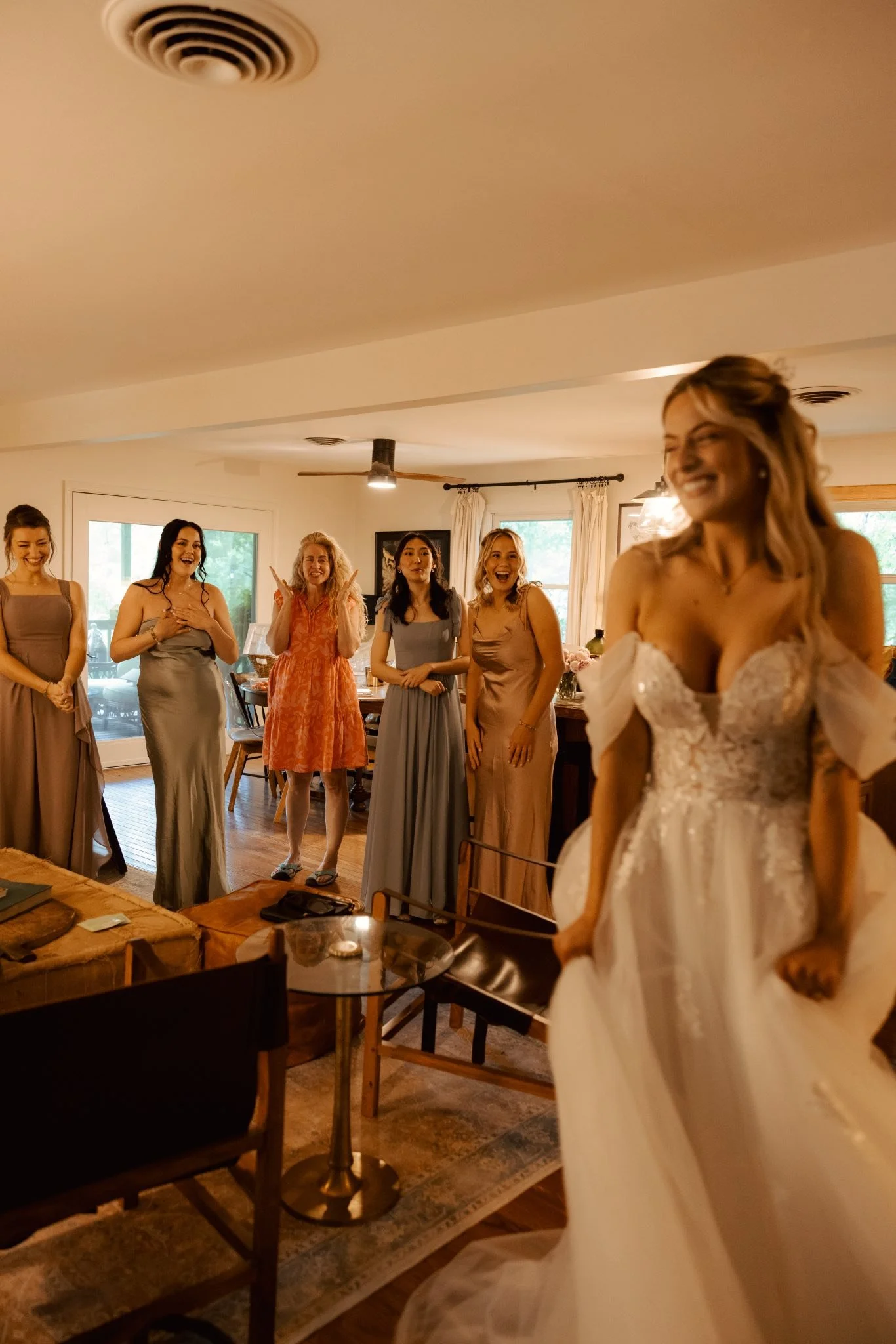 A bride in a white wedding gown is smiling and walking into a room where five women, likely bridesmaids or friends, are standing and reacting happily.