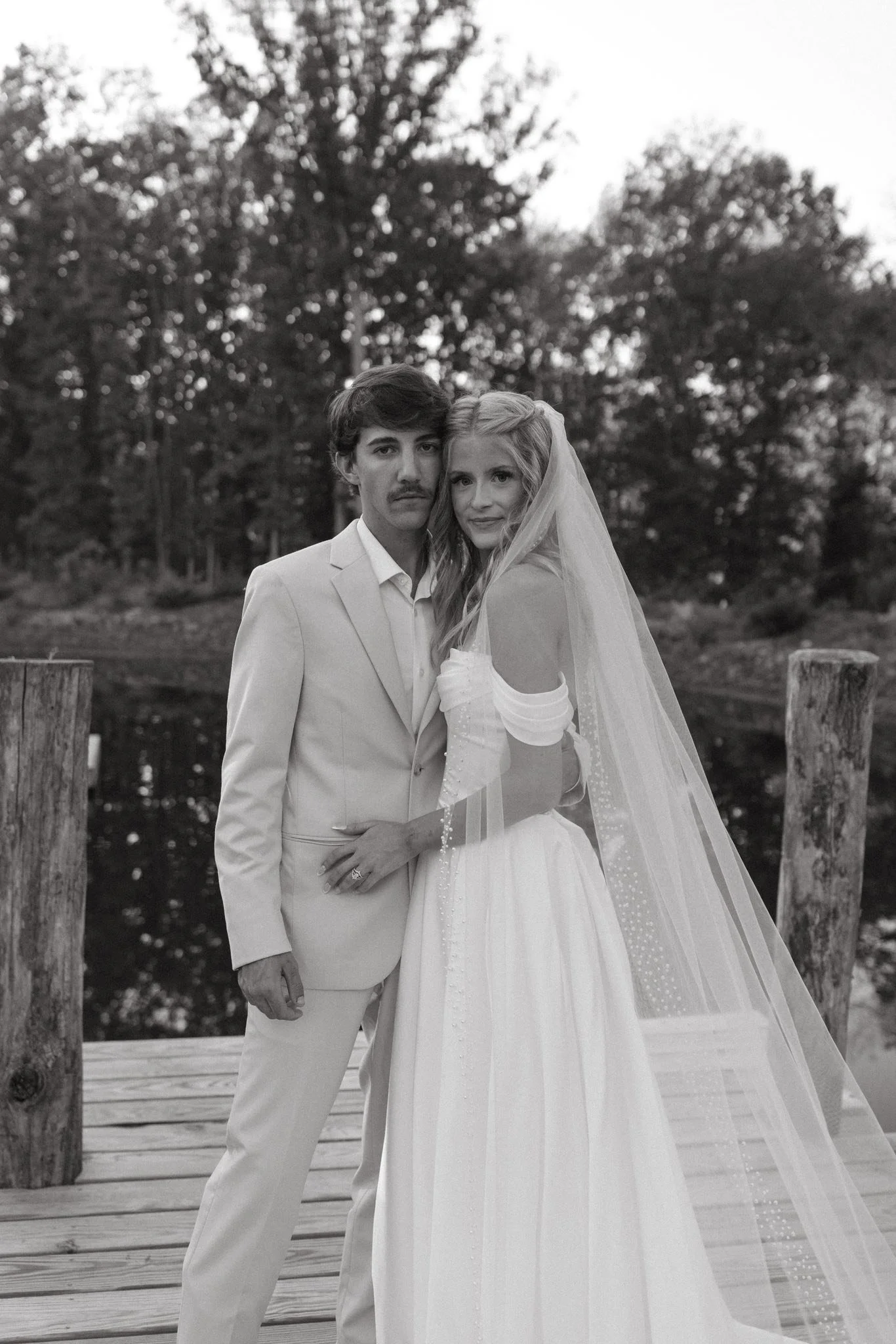 A black and white photograph of a bride and groom standing on a wooden dock near a body of water, with trees in the background. The bride is wearing a wedding dress with a long veil, and the groom is in a light-colored suit.
