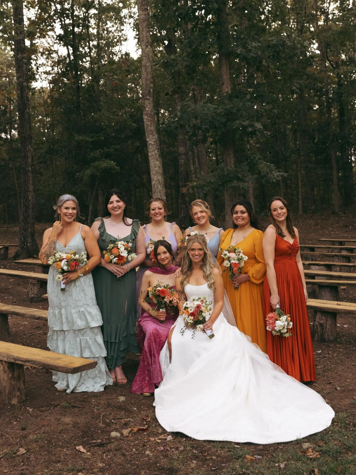 A group of nine women, including a bride in a white wedding dress, posing outdoors in a wooded area, holding bouquets, during a wedding celebration.