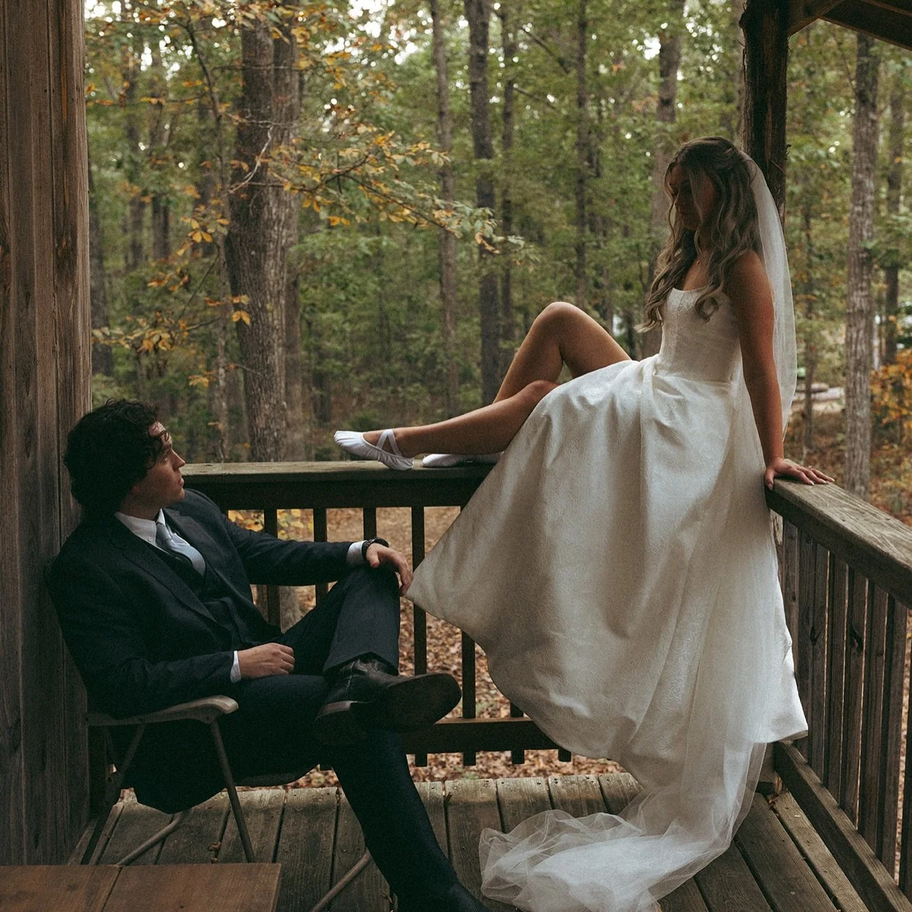 A woman in a wedding dress and veil sitting on a porch railing with one leg crossed over the other, looking down, while a man in a suit and tie sitting in a chair gazes up at her in a wooded outdoor setting.