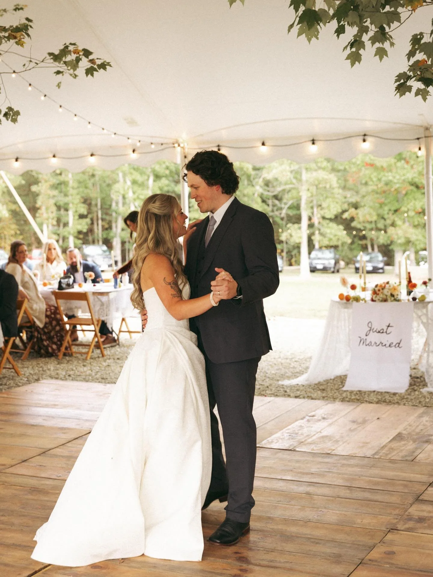 A bride and groom are dancing at their outdoor wedding reception under a white tent with string lights. The bride is wearing a white wedding gown and the groom is in a black suit. There are guests in the background and a sign that reads 'Just Married