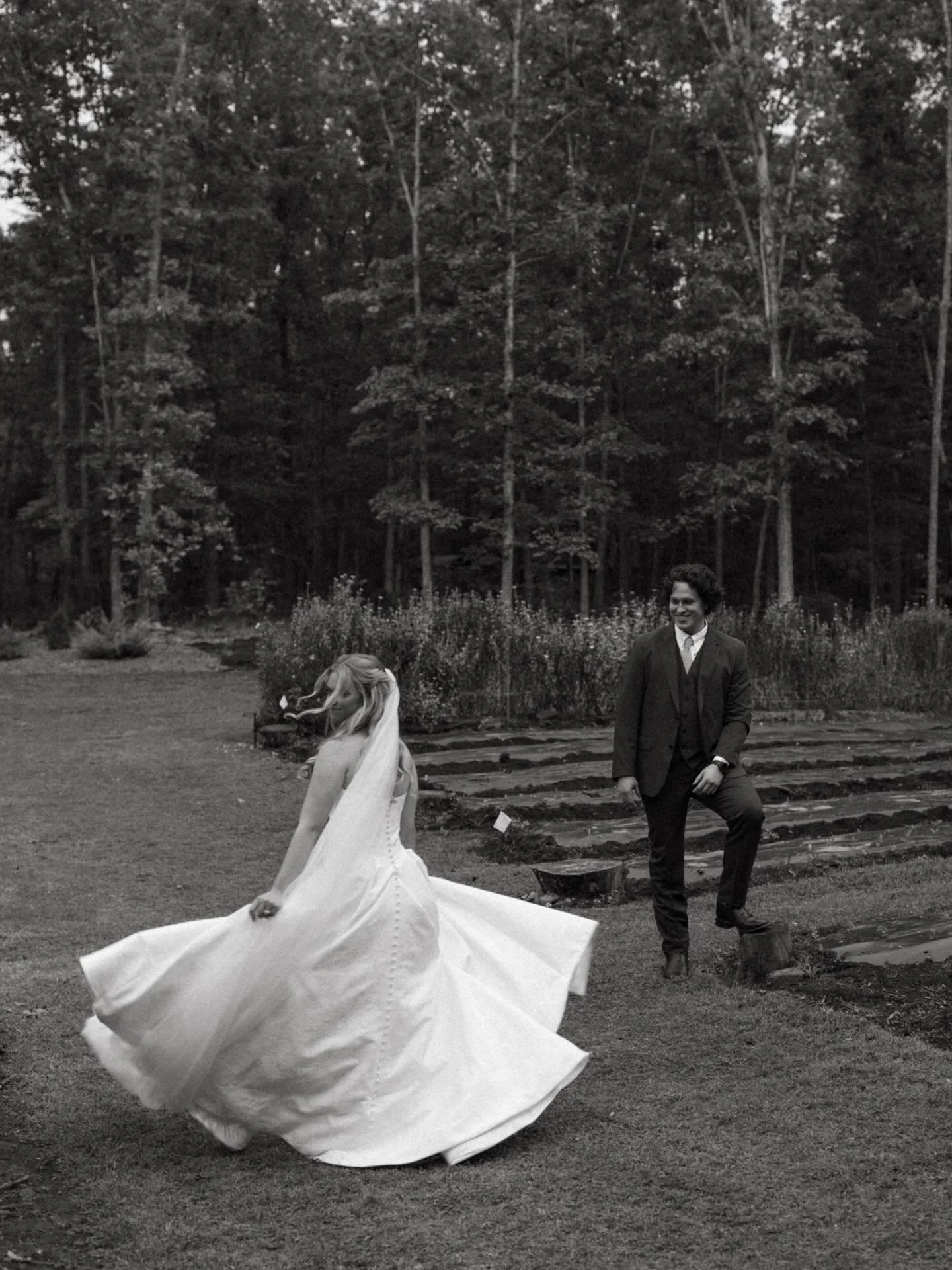 A bride in a wedding dress twirling outdoors with a man in a suit standing nearby, forest background.