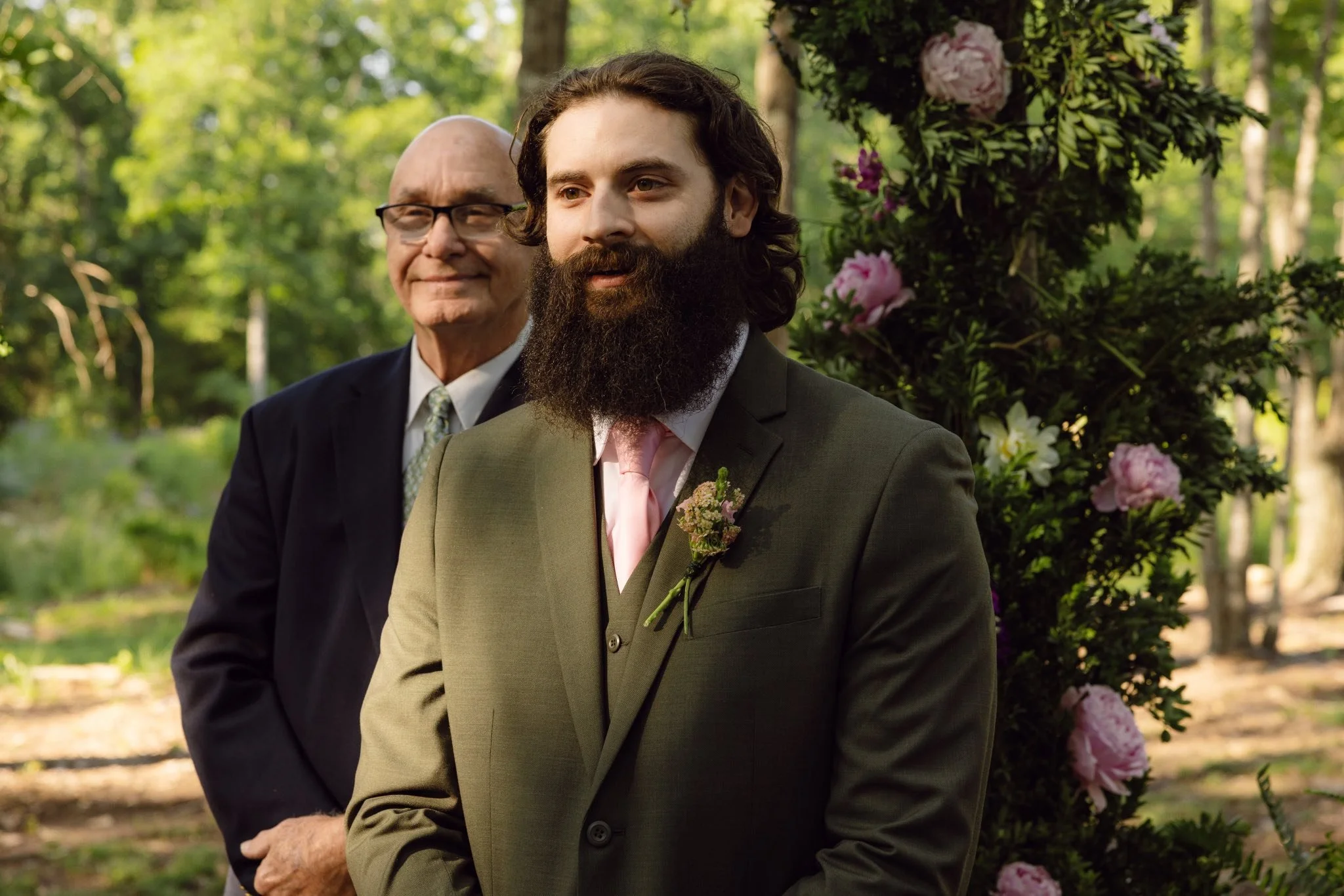 Two men in suits standing outdoors, with one in front having long dark hair and a beard, and the other behind with glasses and a bald head, surrounded by trees and pink flowers.