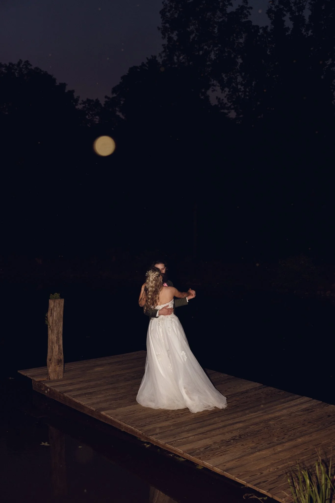 A couple dancing on a wooden dock at night under a full moon, with trees in the background.