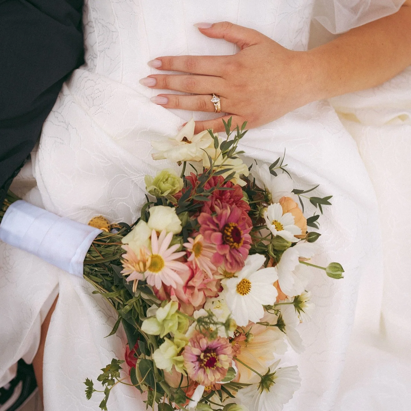 Close-up of a bride's hand with an engagement ring, resting on her white wedding dress, with a colorful bouquet of flowers.