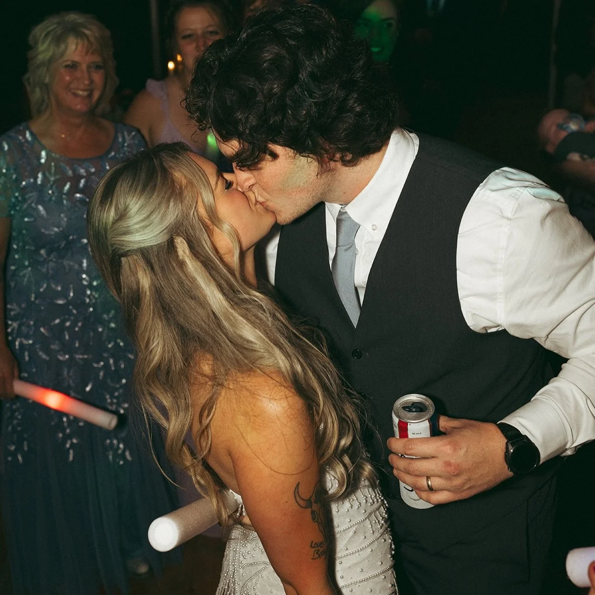 A couple sharing a kiss at a celebration or wedding, with guests in the background smiling and enjoying the moment.