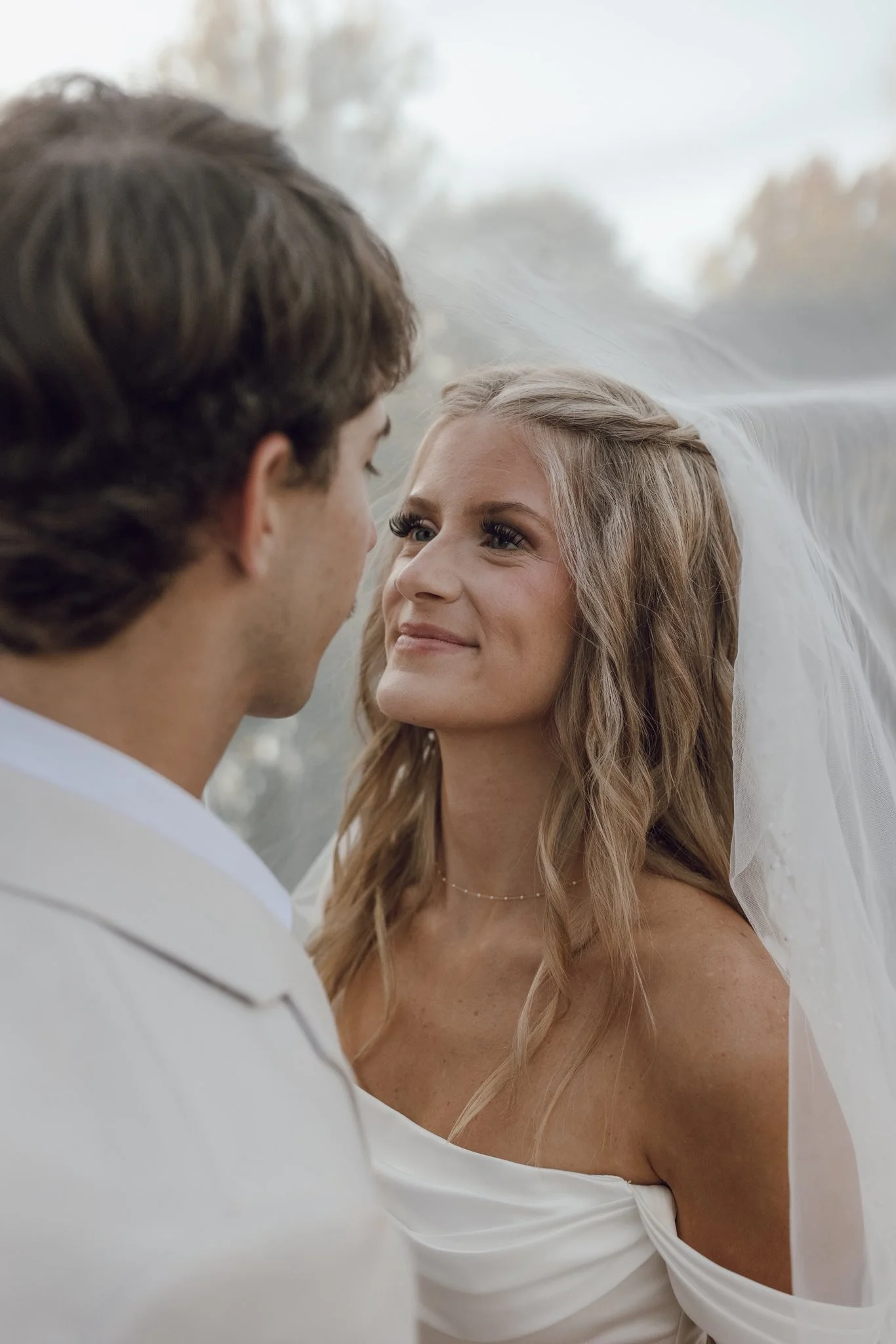 A bride and groom facing each other outdoors, close-up, with the bride smiling and the groom's face partially visible.