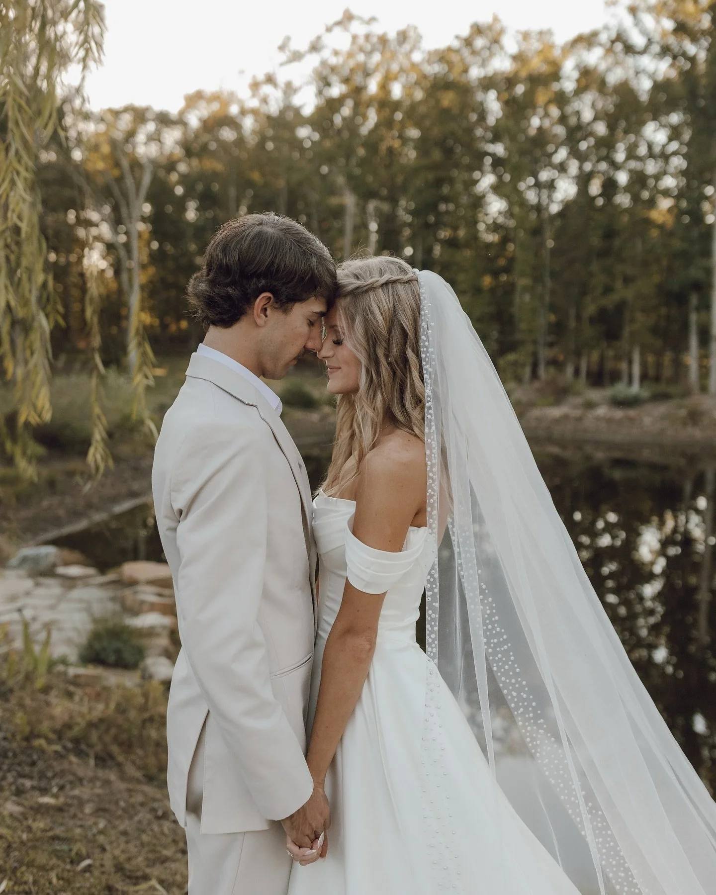 A bride and groom stand close together outdoors, with foreheads touching and eyes closed, holding hands during their wedding.