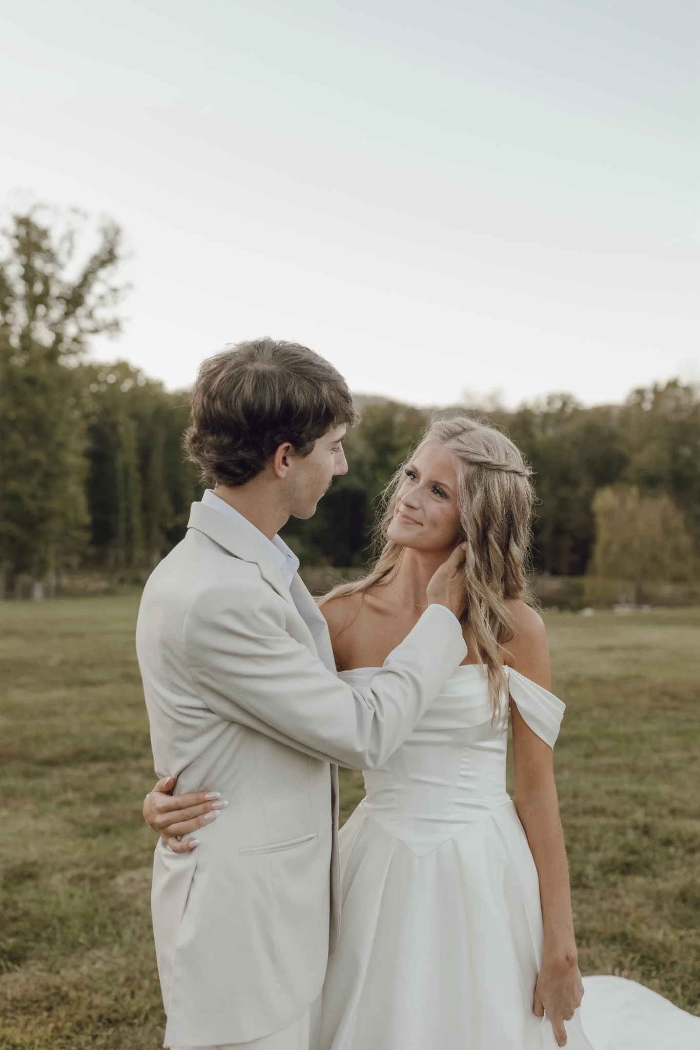 A man and woman in wedding attire standing outdoors on a grassy field with trees in the background, gazing at each other.