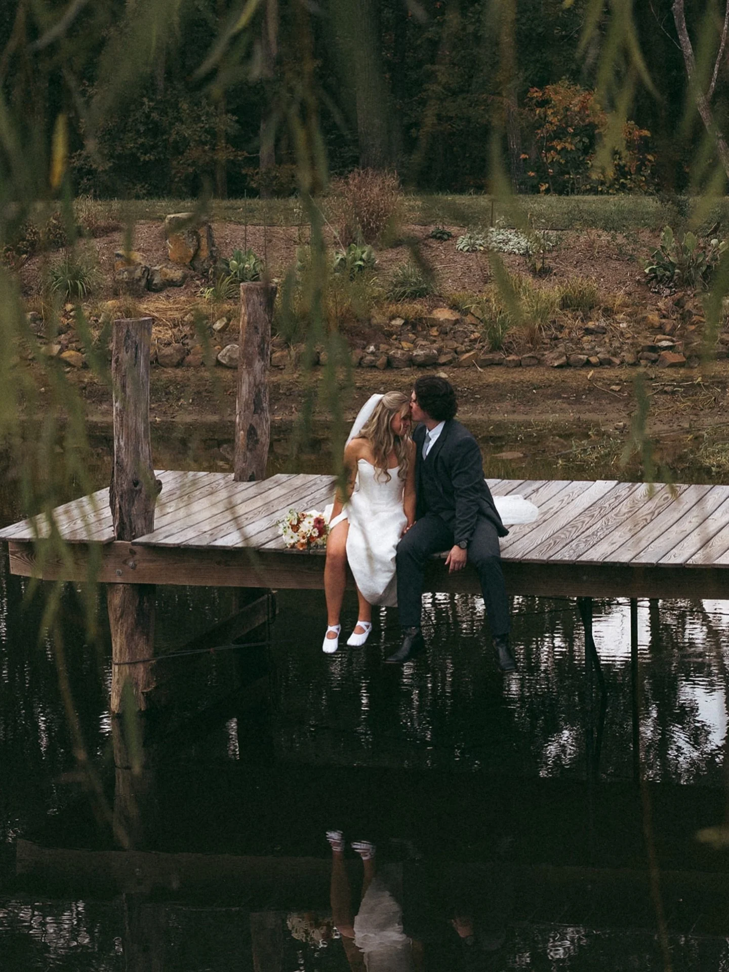 A bride and groom sitting on a wooden dock by a pond, sharing an intimate moment, surrounded by natural greenery and landscape.