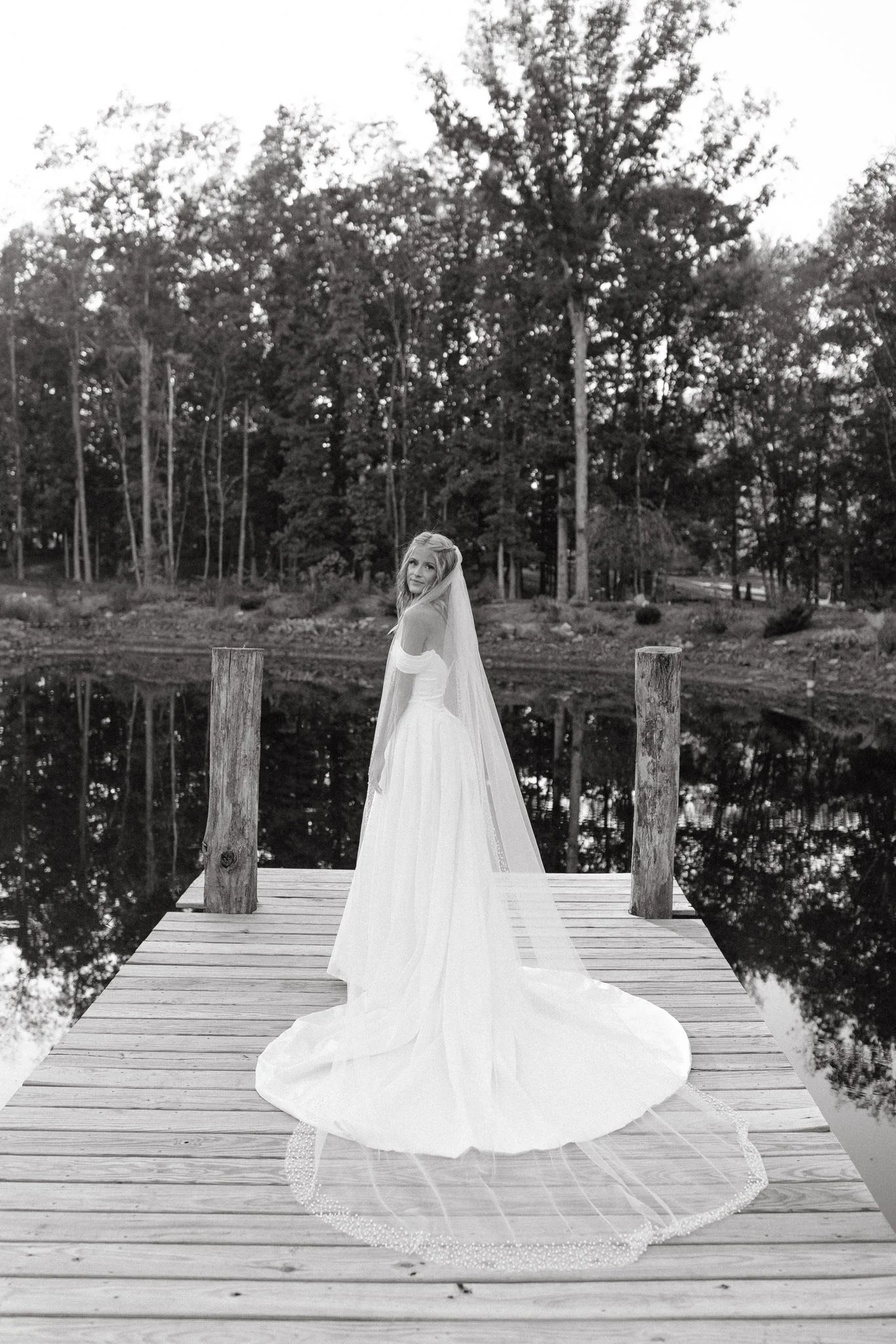 A woman in a wedding dress standing on a wooden dock by a pond, surrounded by trees, in a black and white photo.